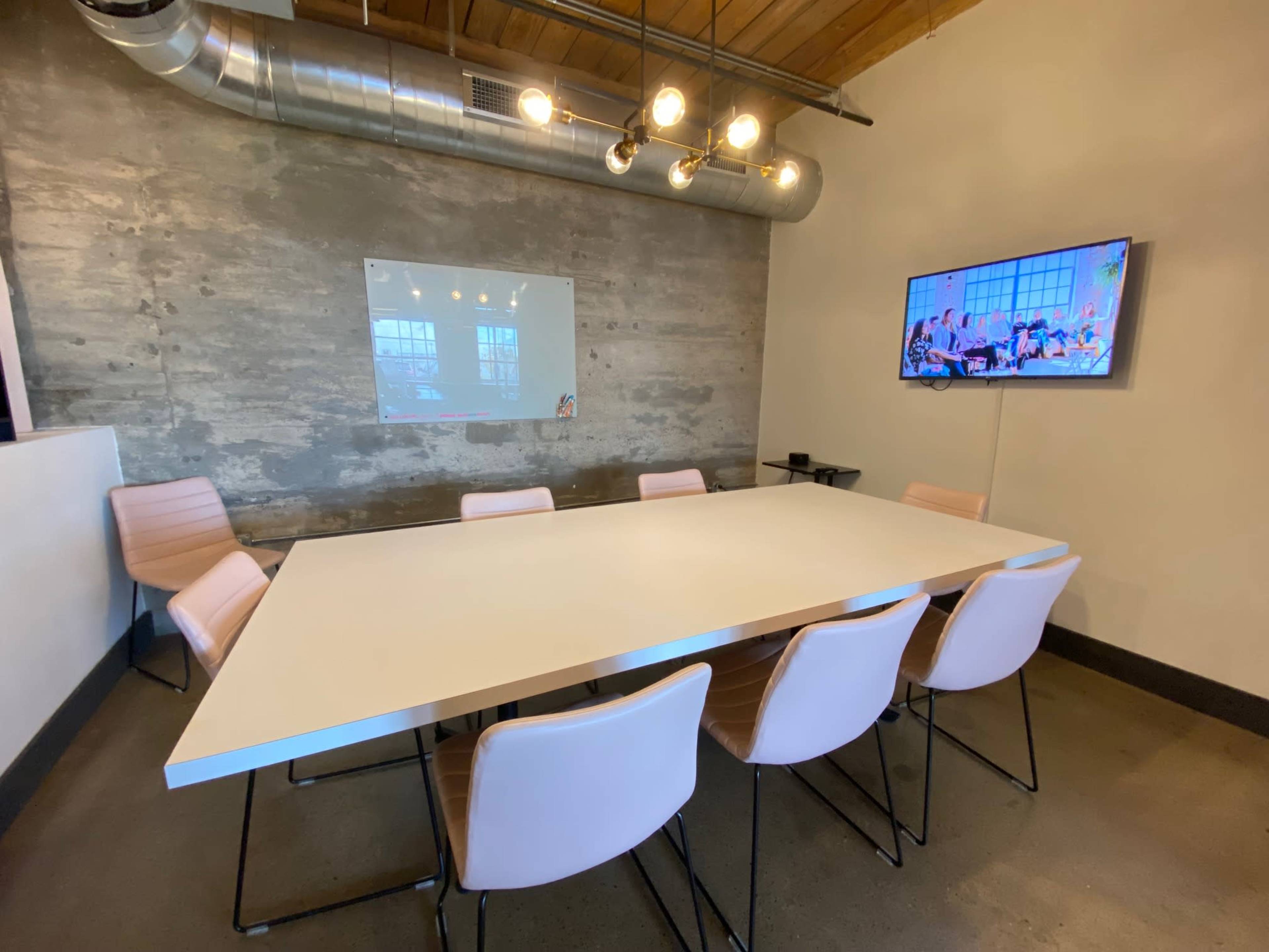 A modern conference room features a long white table surrounded by pink chairs, with a wall-mounted TV and a whiteboard.