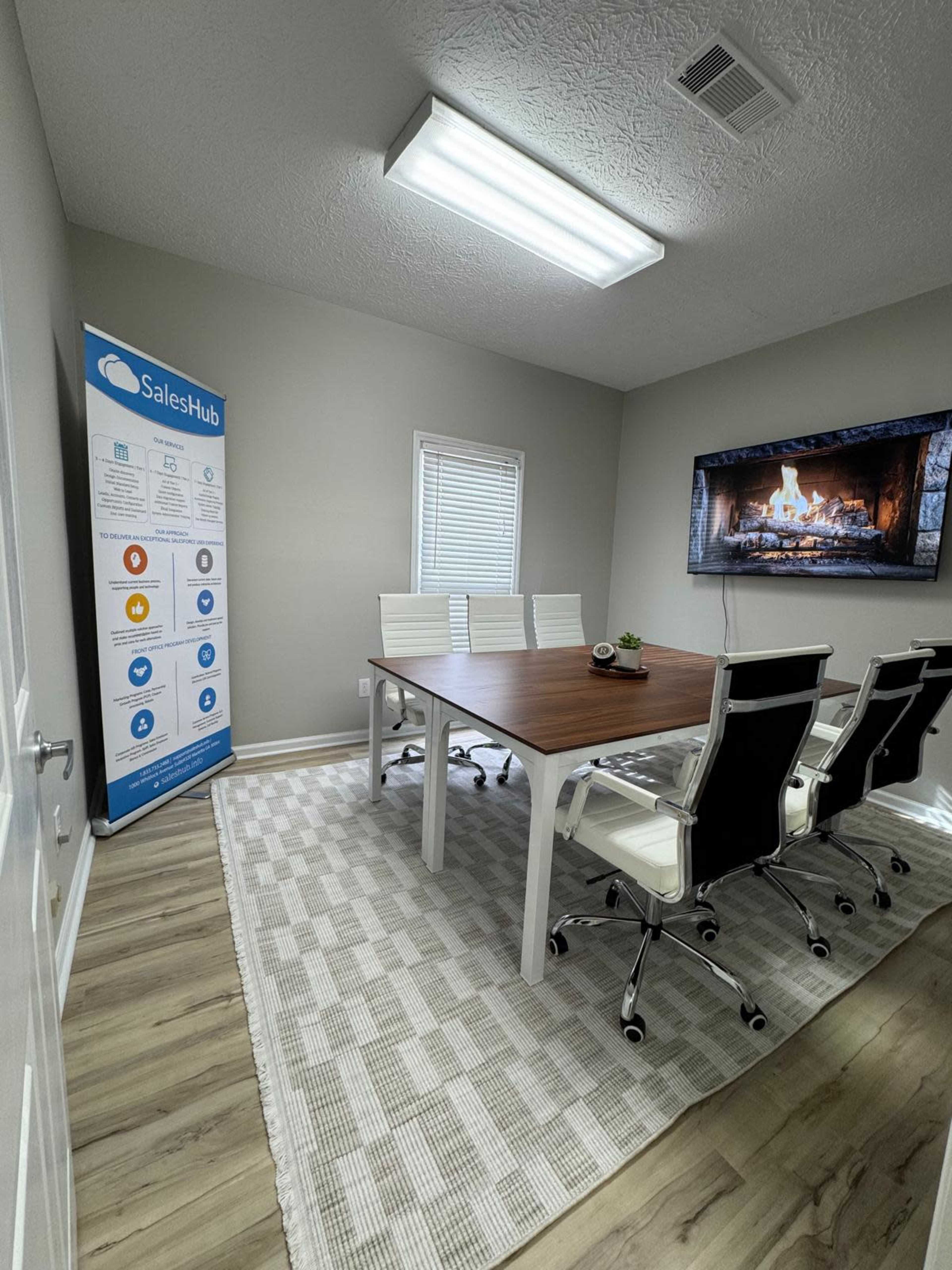 The image shows a modern conference room with a large wooden table surrounded by white office chairs, a banner displaying information about SalesHub on the wall, and a television featuring a fireplace display.