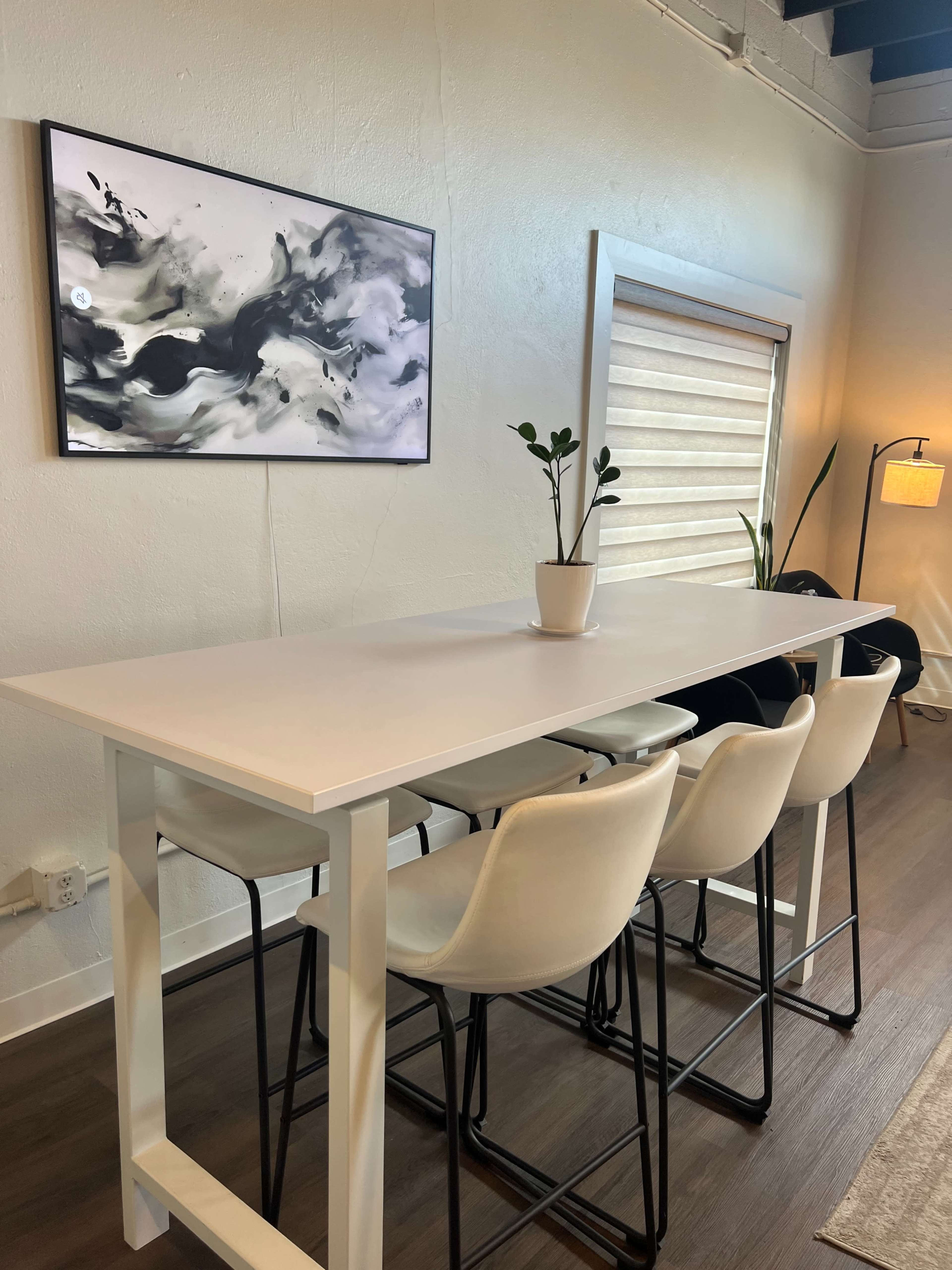 A modern dining area features a tall white table with four white chairs, a potted plant in the center, and a large abstract painting on the wall.