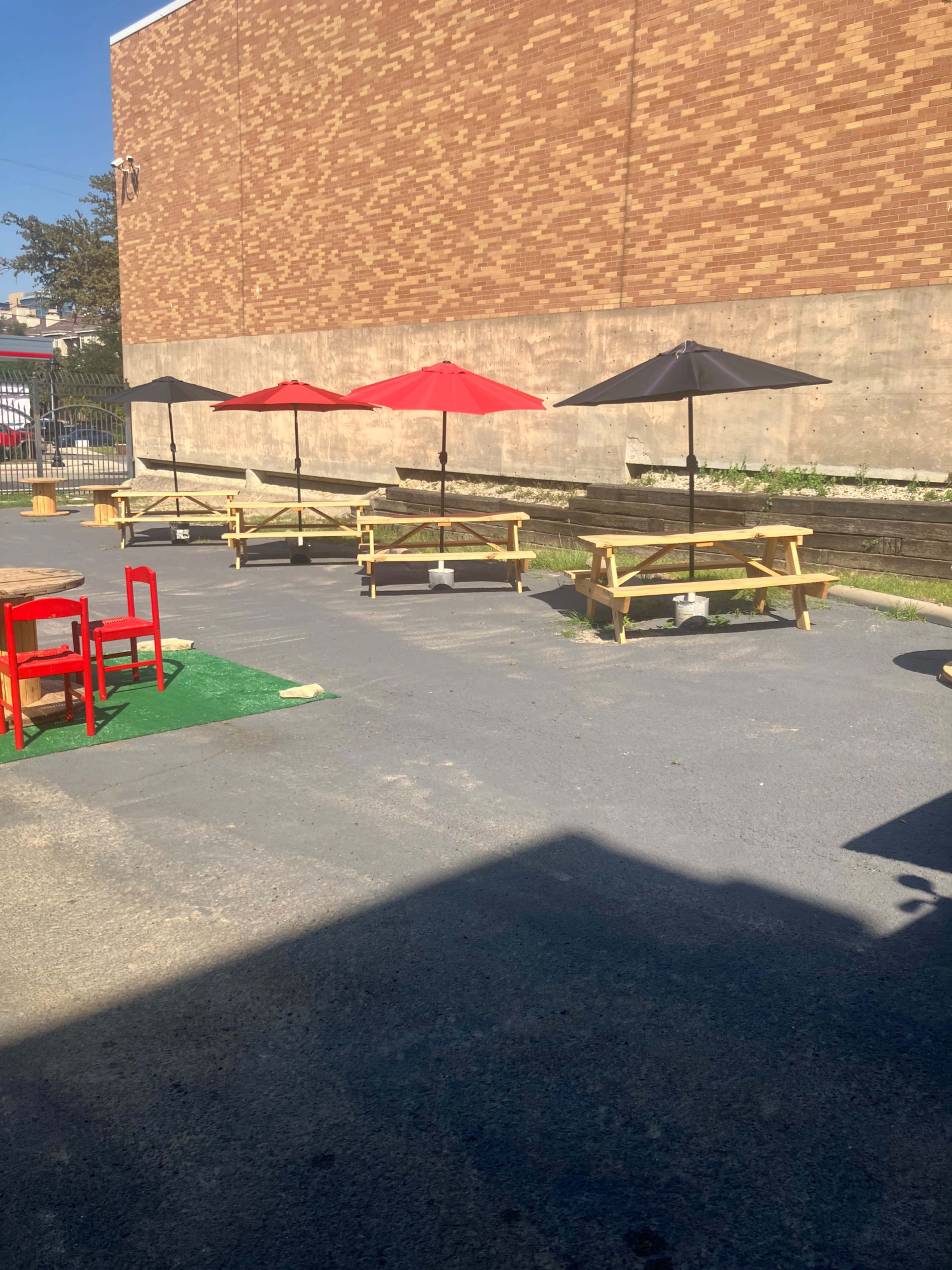 The image shows an outdoor seating area with picnic tables and umbrellas in shades of red and black, set against a brick wall.