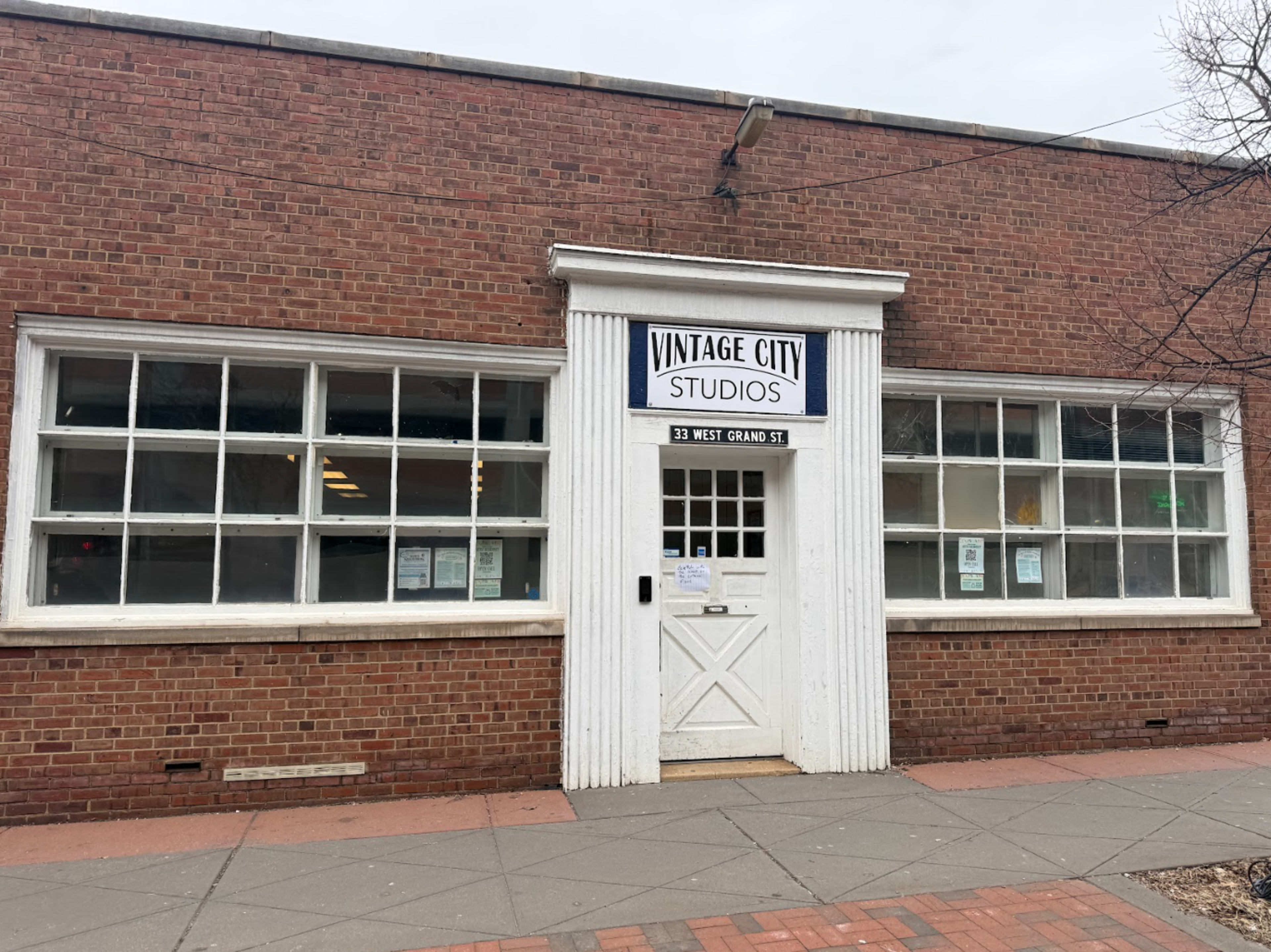 The image shows the façade of a brick building with large windows and a sign reading "Vintage City Studios" above the entrance door.