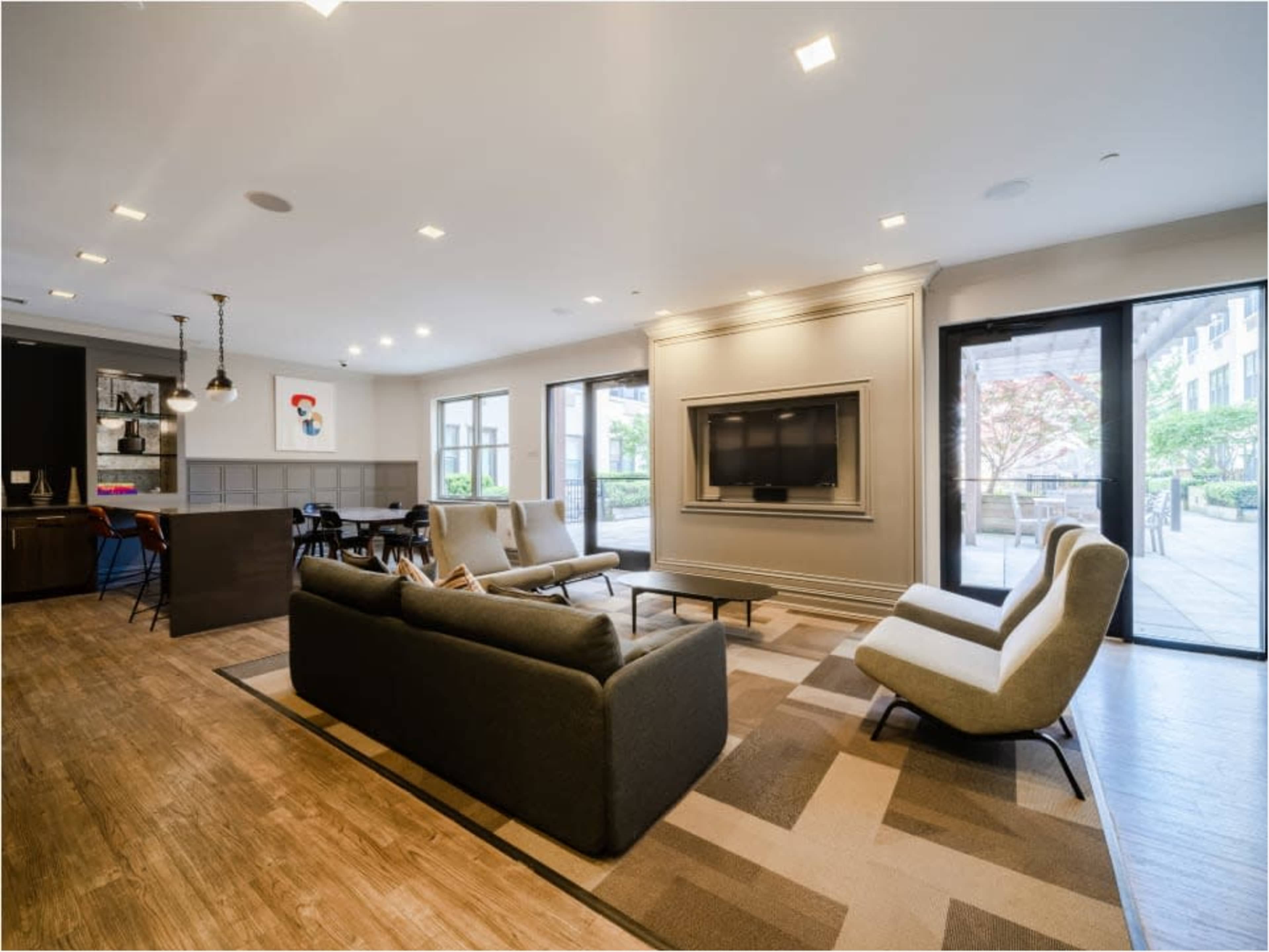 The image shows a modern living room with a gray sofa, two light chairs, a television mounted on the wall, and large windows that provide natural light.