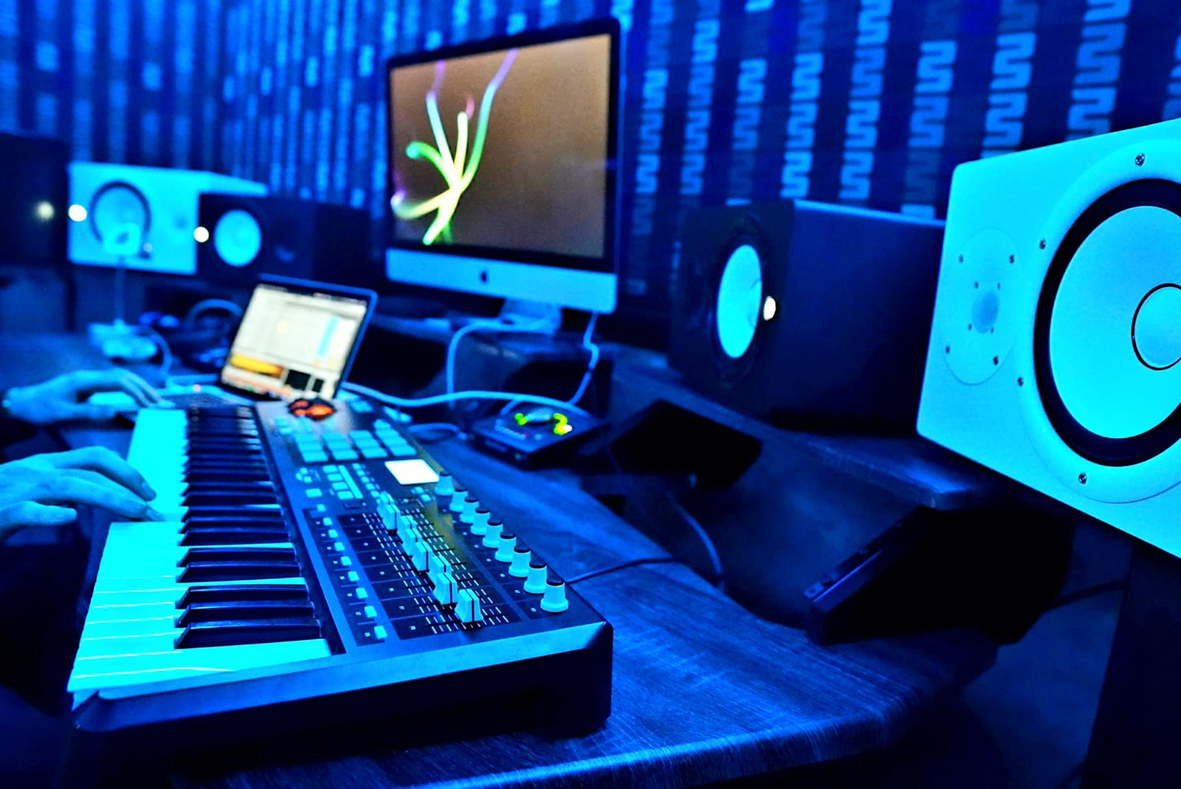 A person plays a keyboard in a dimly lit music studio with a computer and studio monitors in the background.