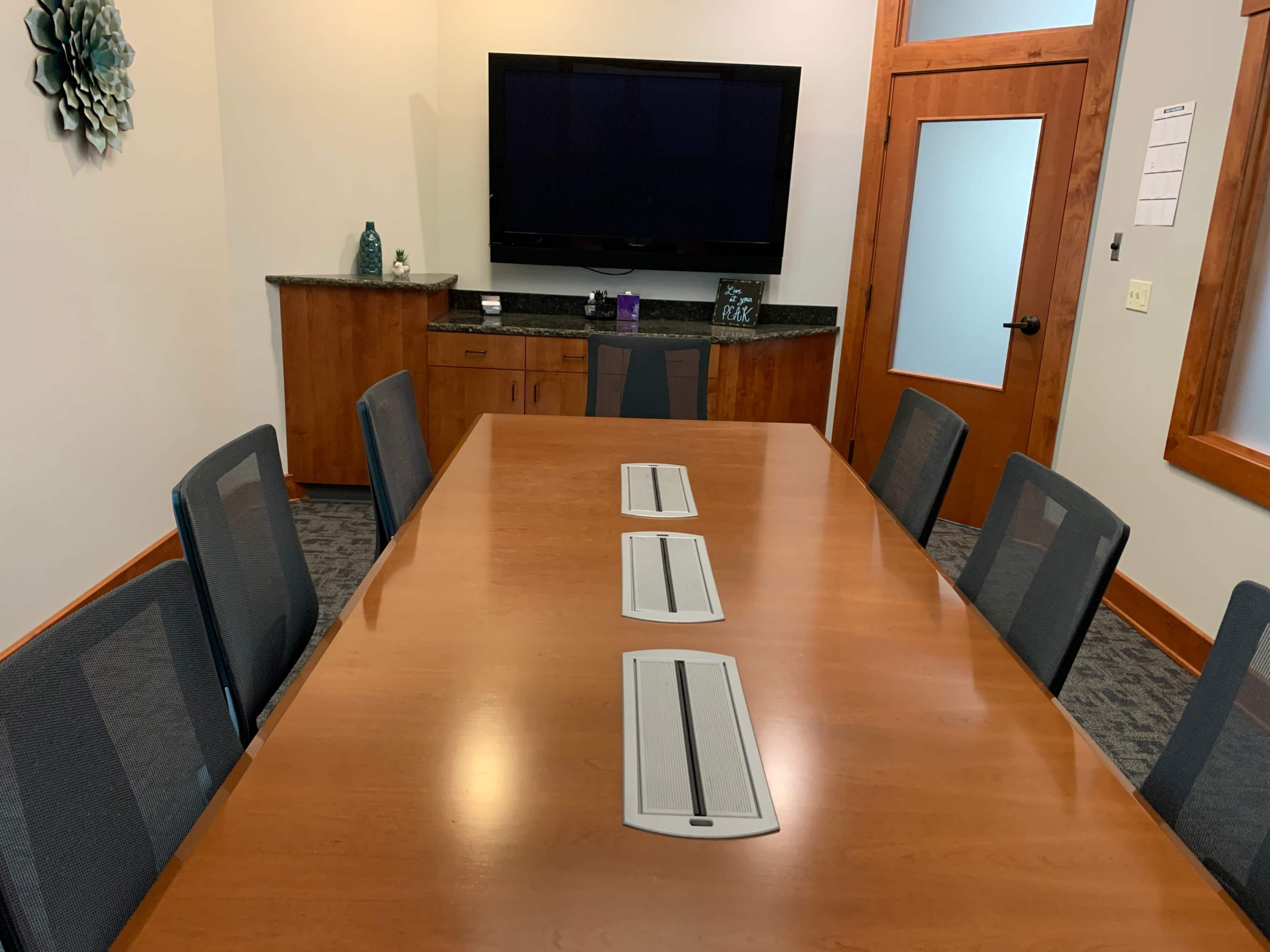 A conference room features a wooden table with three cable management ports, surrounded by six chairs and equipped with a television mounted on the wall.