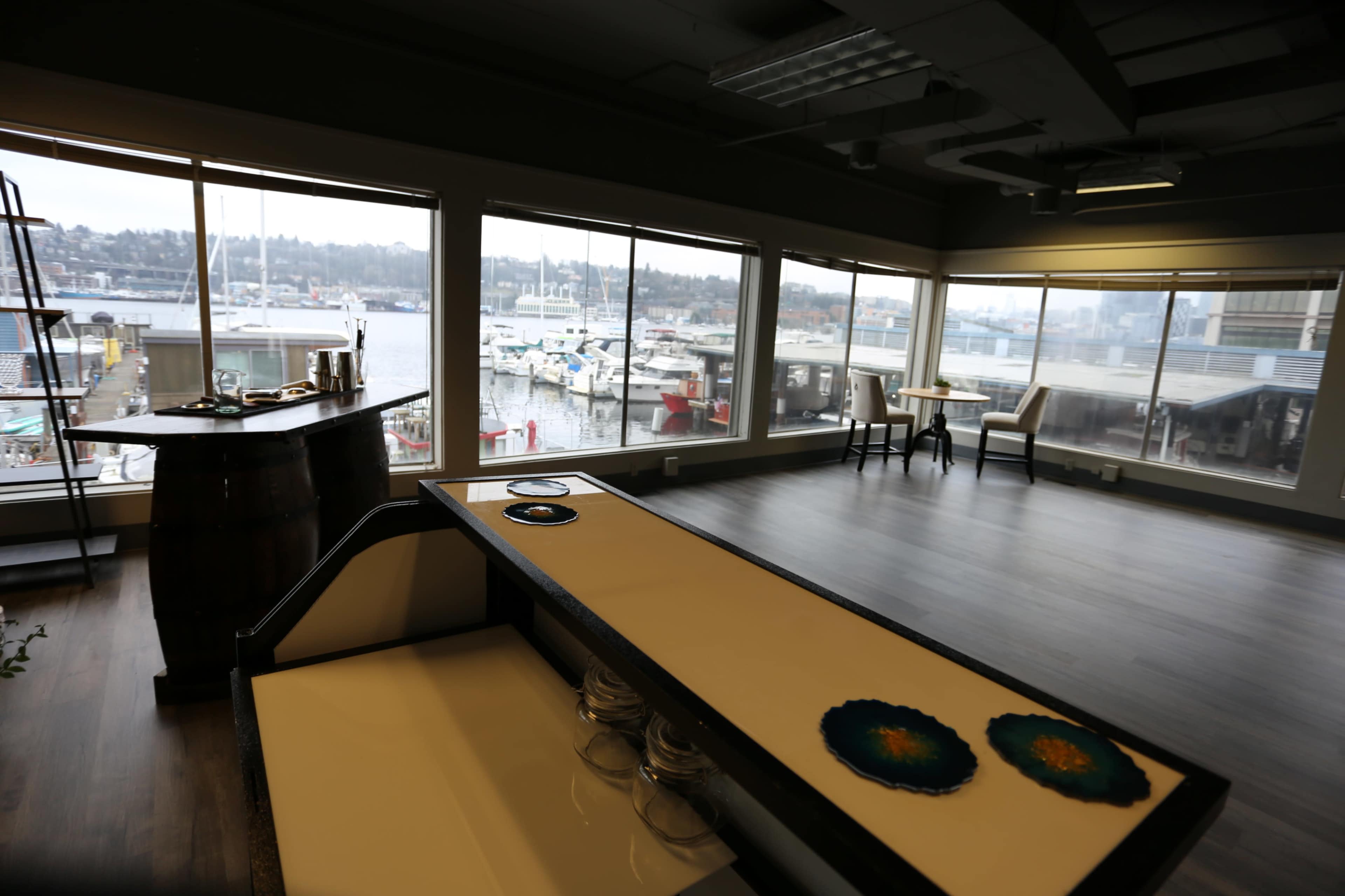 The interior of a waterfront venue features a large window overlooking a marina with various boats, and tables arranged for dining.