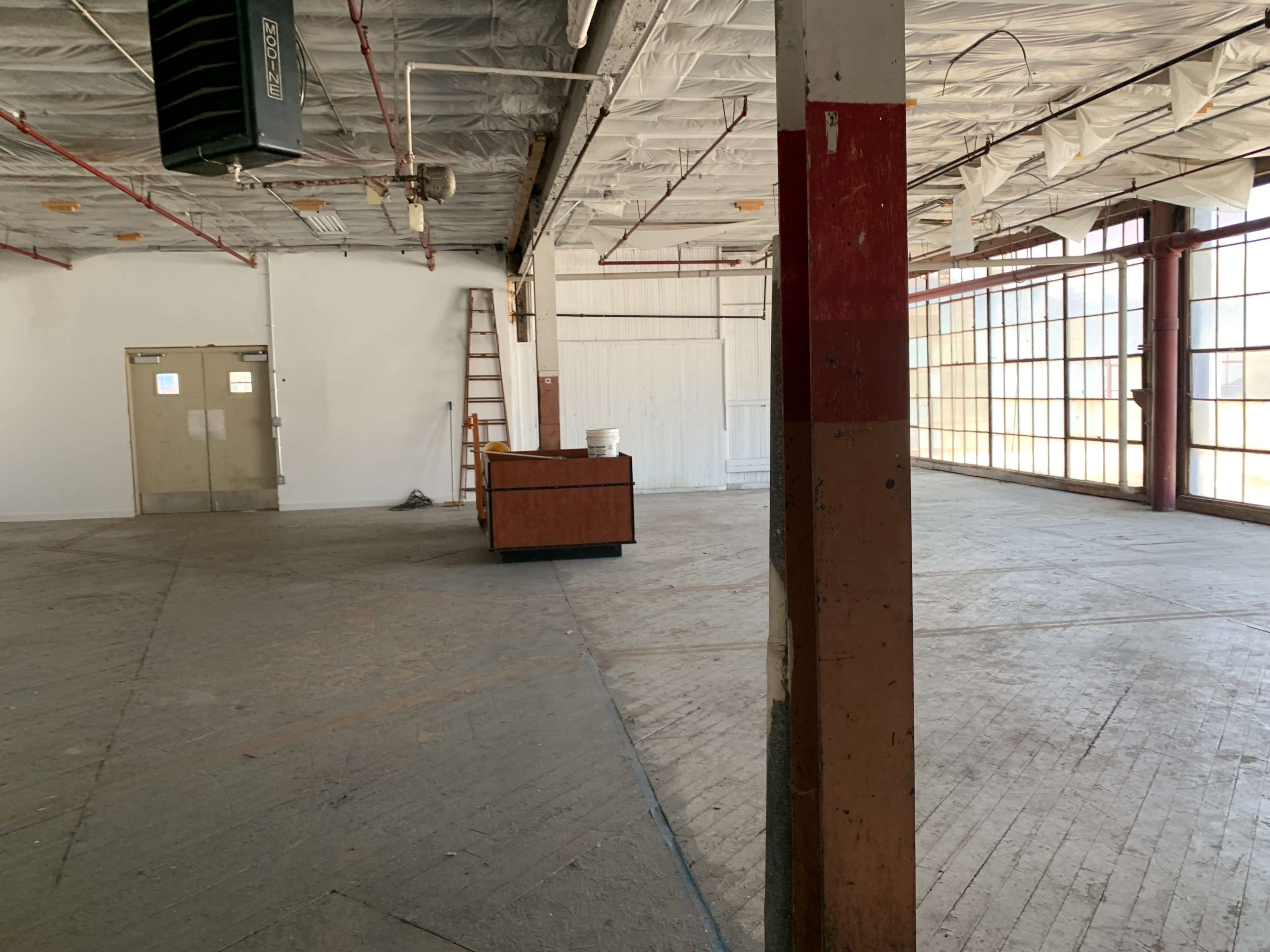 The image shows an empty industrial space with a wooden counter in the center, large windows along one wall, and a ladder against a back wall.