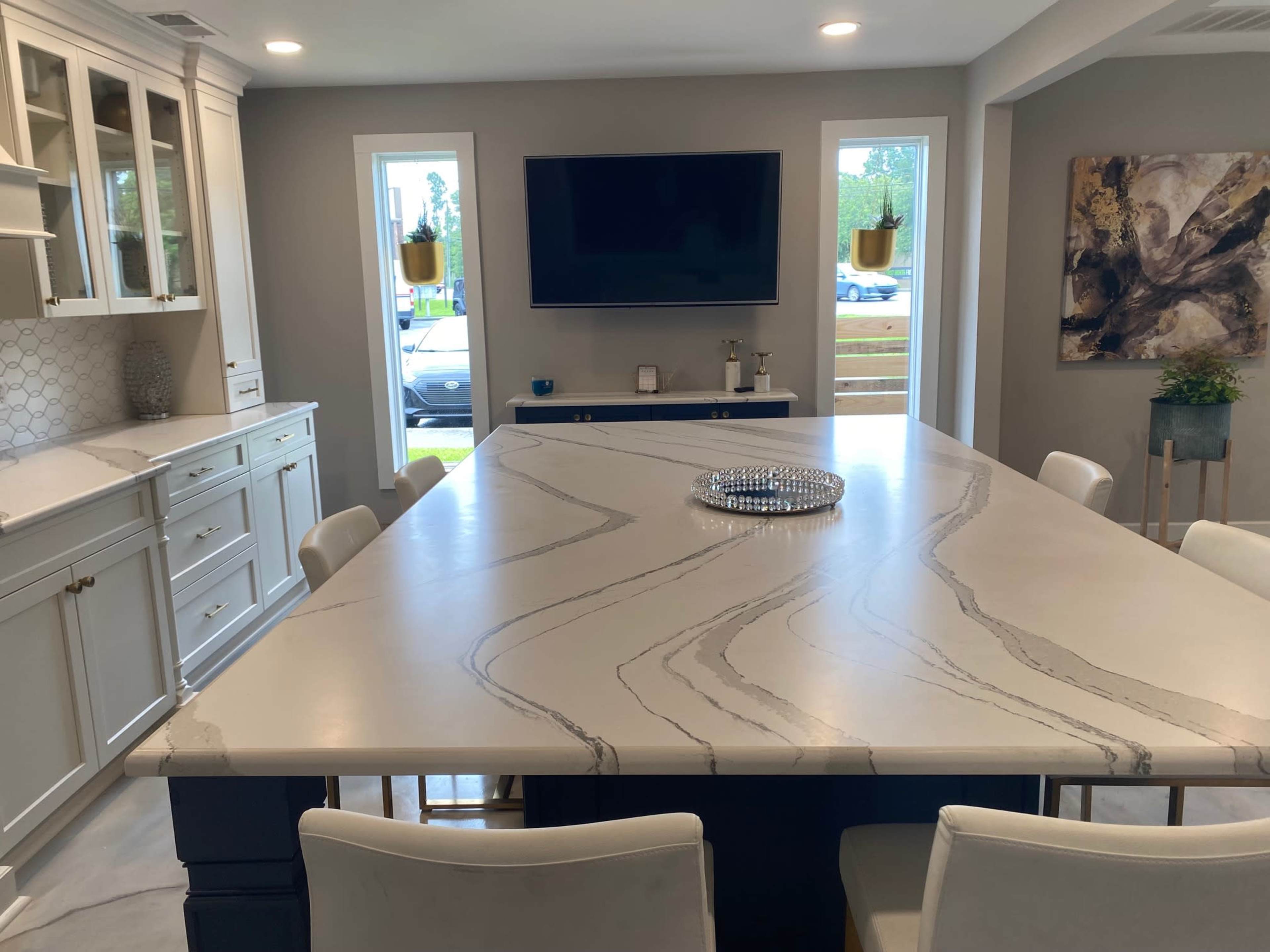 A modern kitchen with a large marble island, white cabinetry, and a flat-screen TV mounted on the wall.