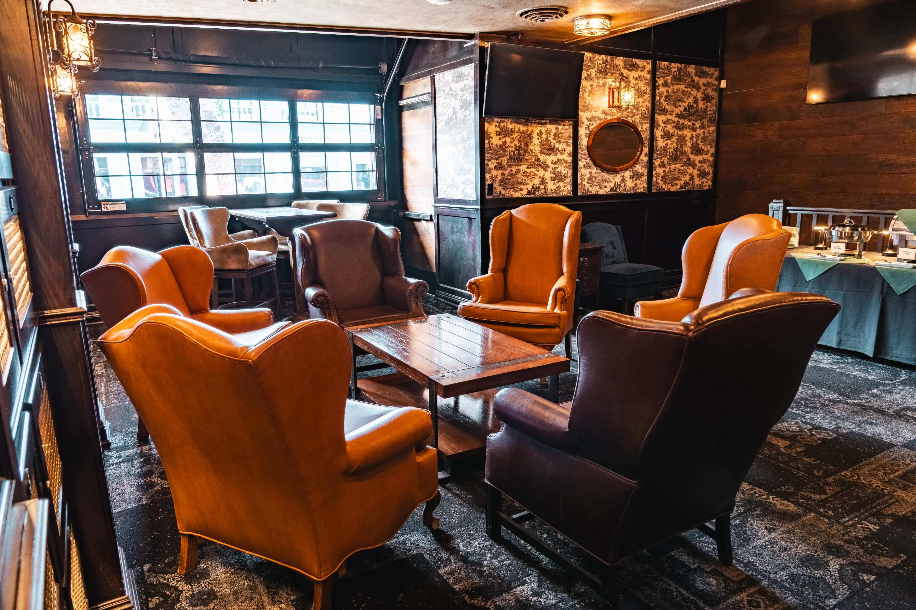 A cozy seating area in a dimly lit lounge features several brown leather wingback chairs arranged around a wooden coffee table.