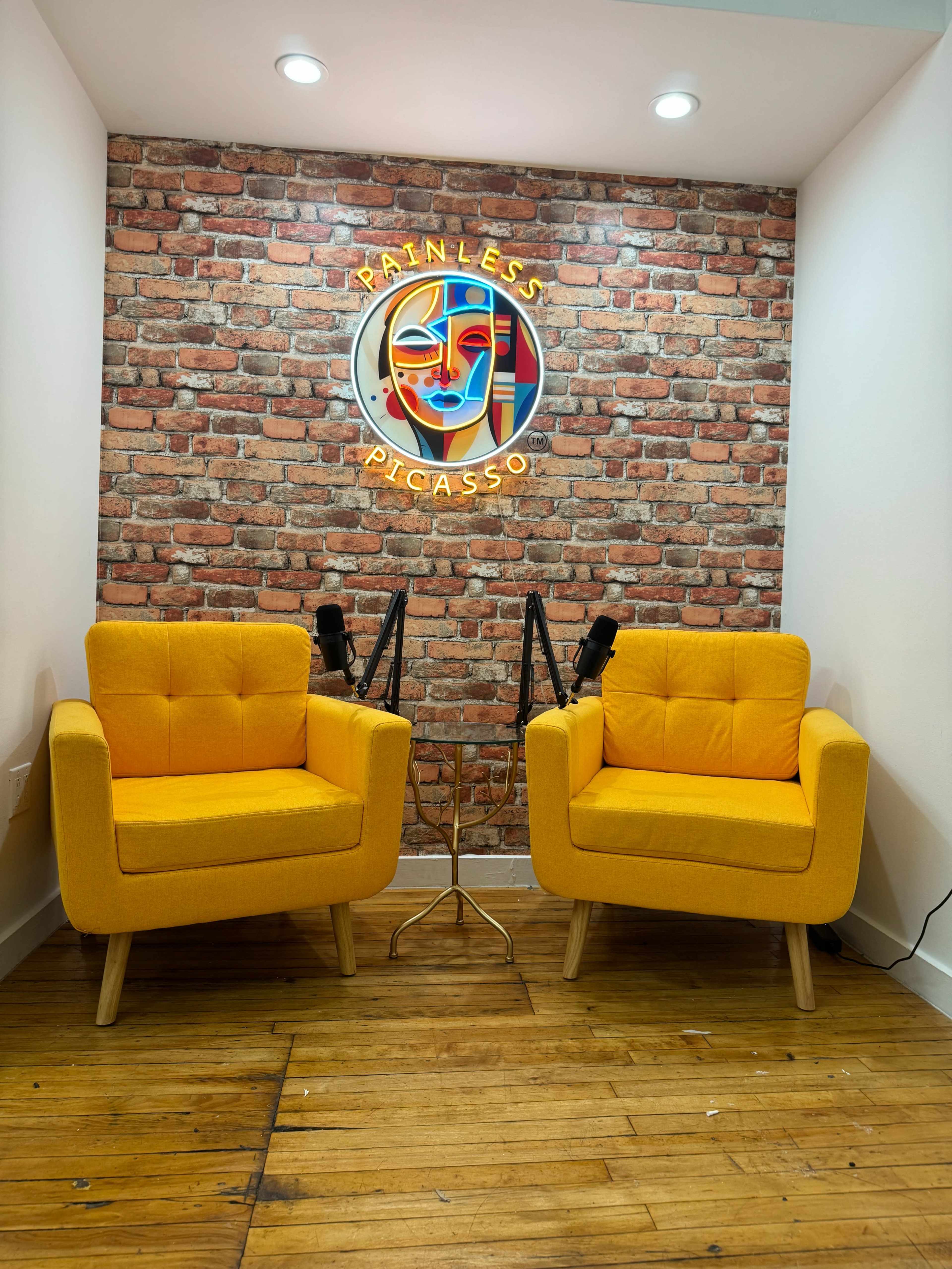 The image shows a small recording studio with two yellow armchairs, a small round table, and a brick-patterned wall featuring a circular logo with the words "Painless Picasso."
