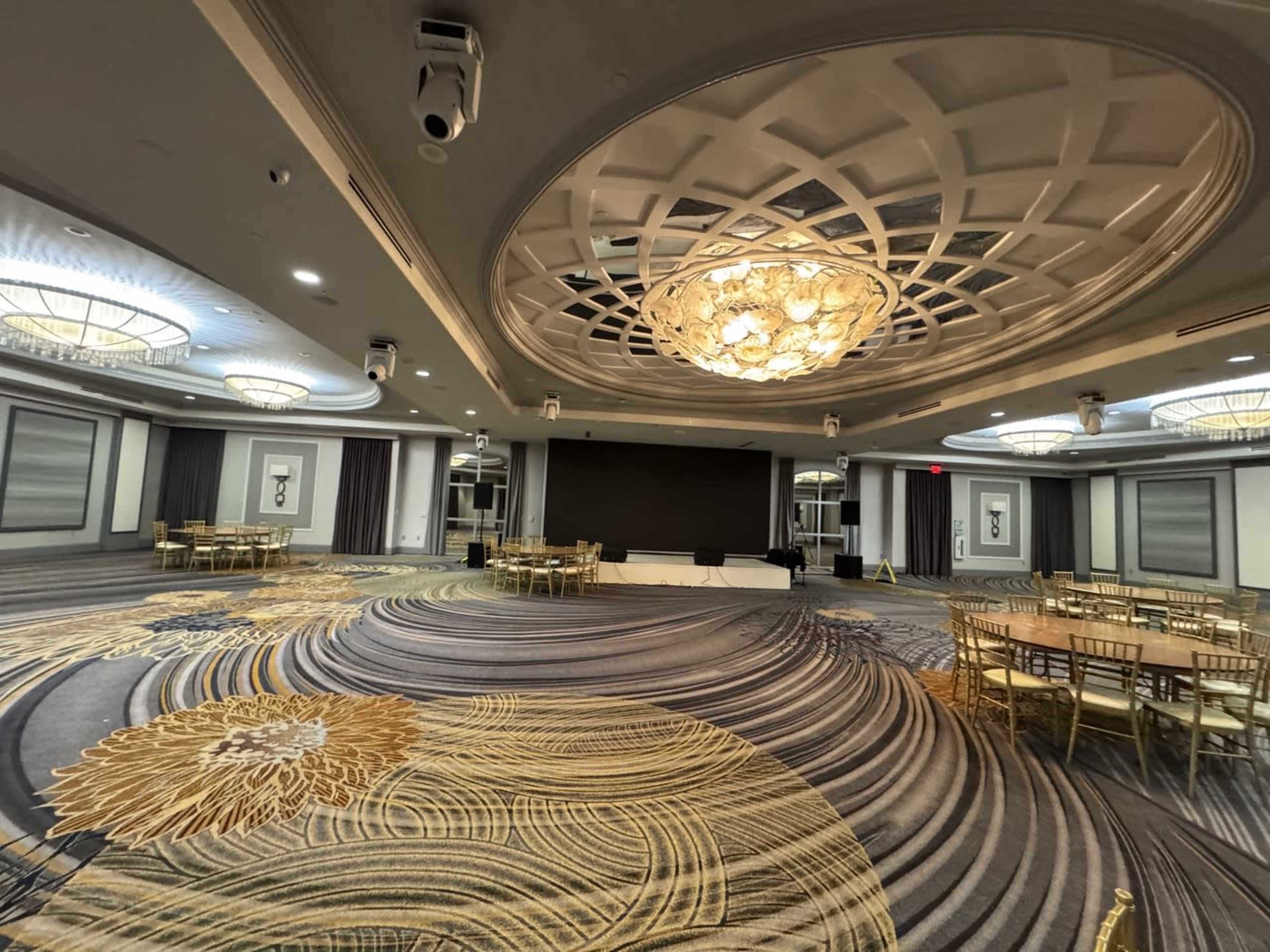 A spacious banquet hall features intricate carpet patterns, a large chandelier, and several round tables set up for an event.