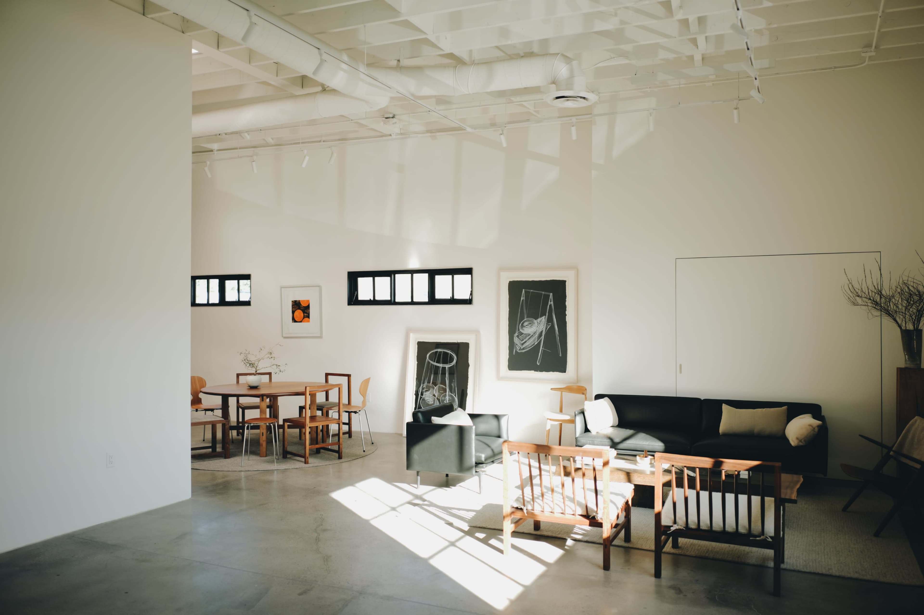 A modern interior space features a seating area with black and wooden furniture, framed artwork on the walls, and a dining area with wooden tables and chairs.