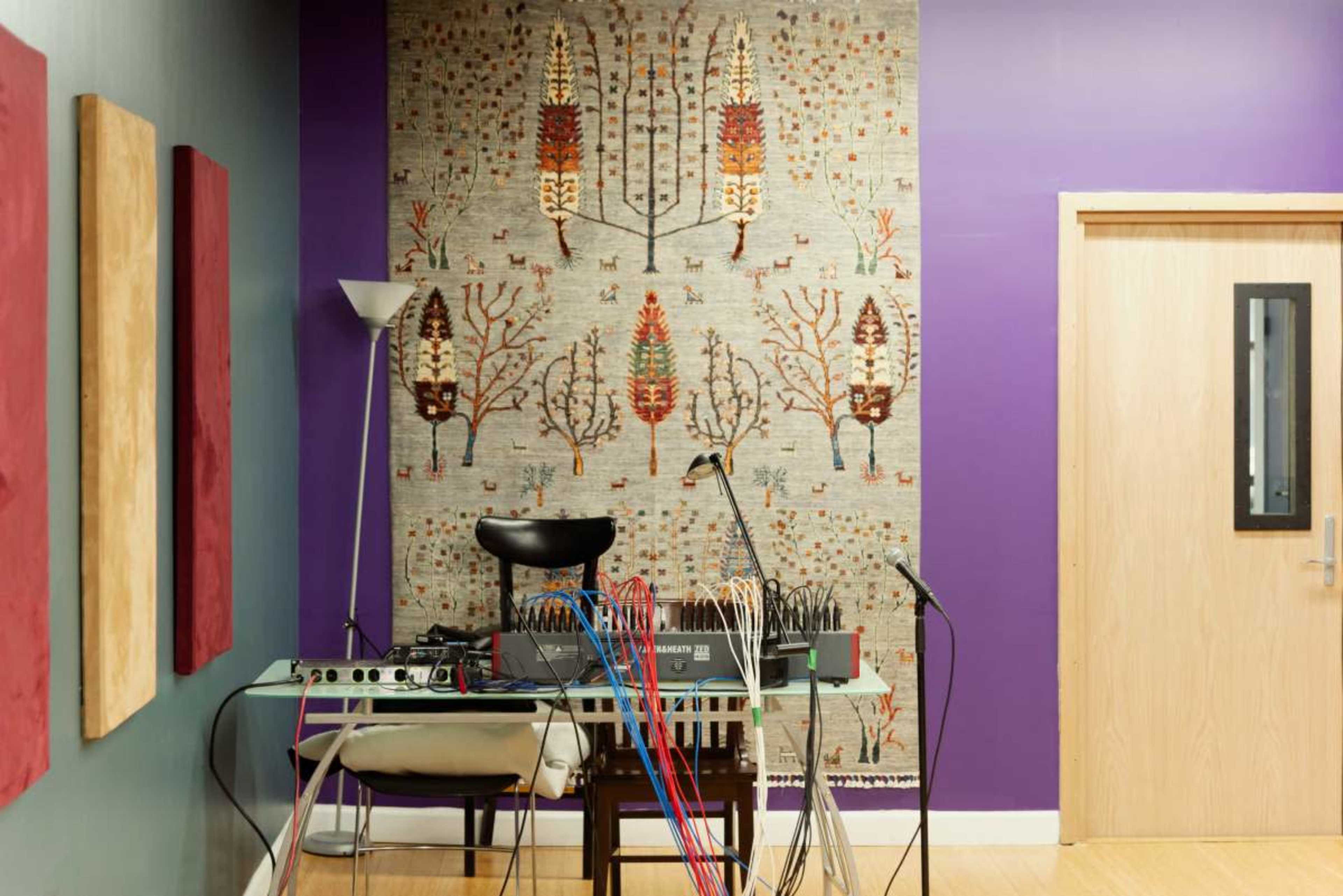 The image shows a corner of a room with a decorated wall featuring a patterned tapestry, a table with electronic musical equipment, and colorful cables.
