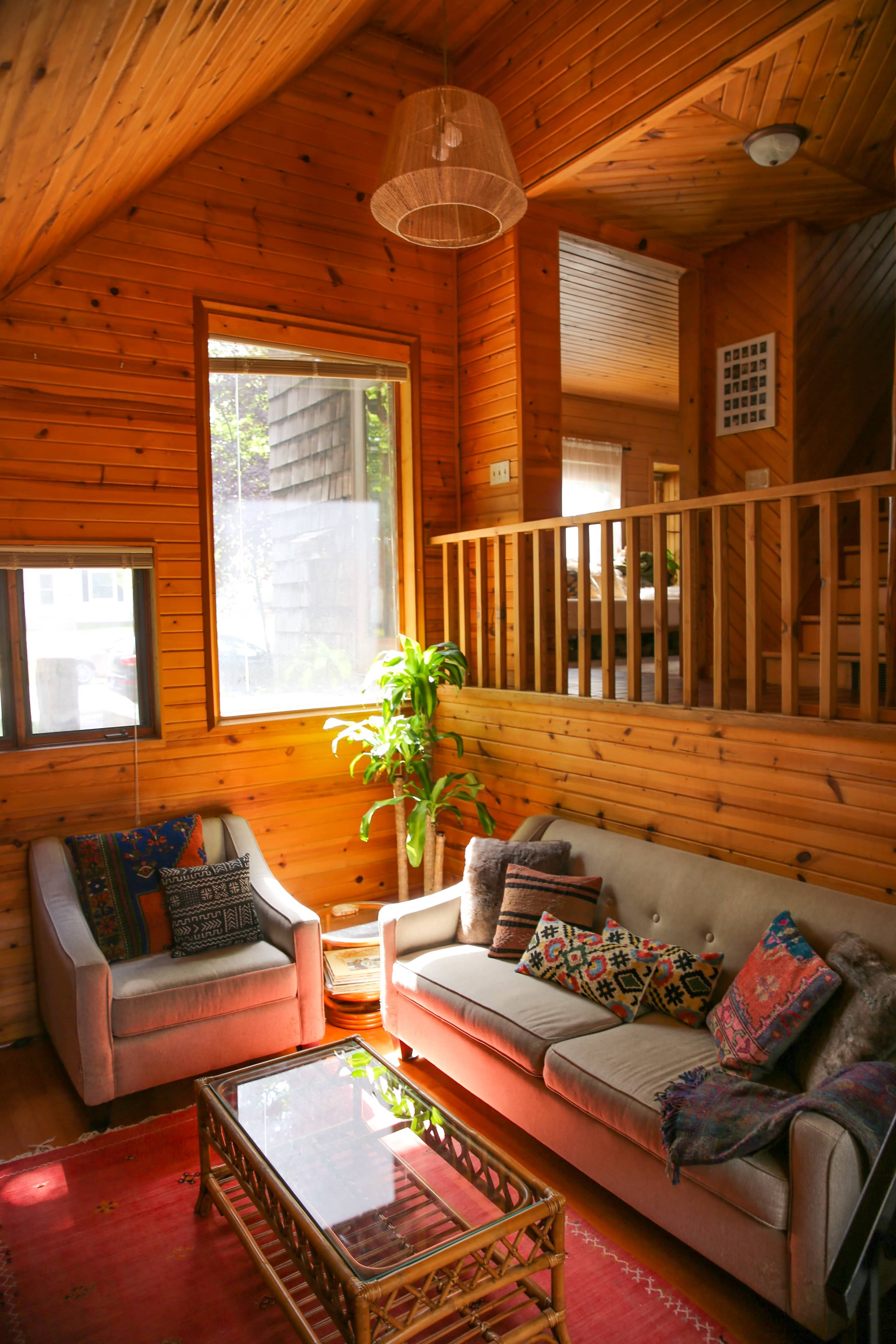 A cozy living room with wooden walls, a gray couch adorned with colorful pillows, a glass coffee table, and a potted plant near a large window.