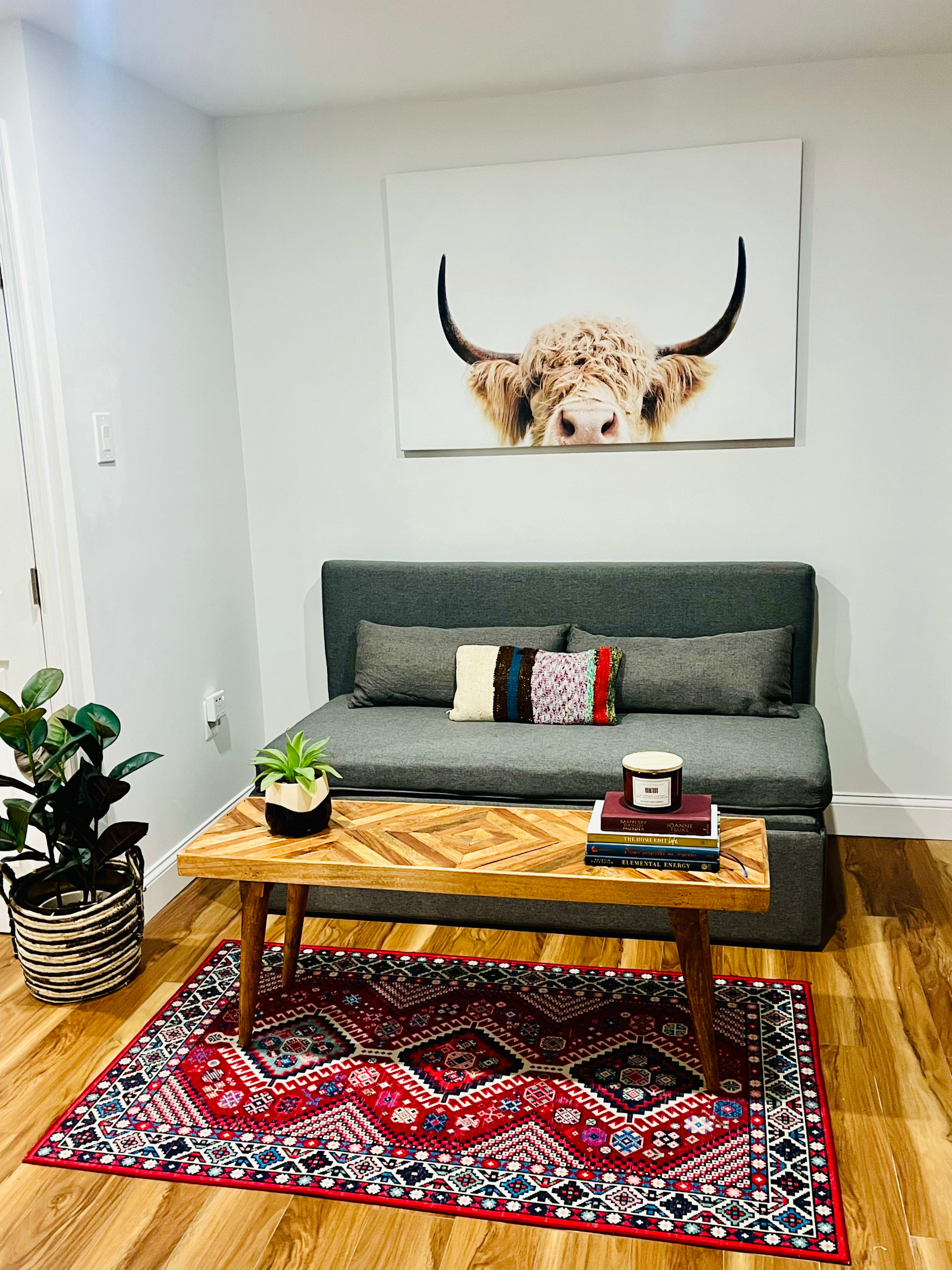A cozy living area features a gray loveseat, a wooden coffee table with books and a candle, a colorful area rug, and a framed picture of a highland cow on the wall.