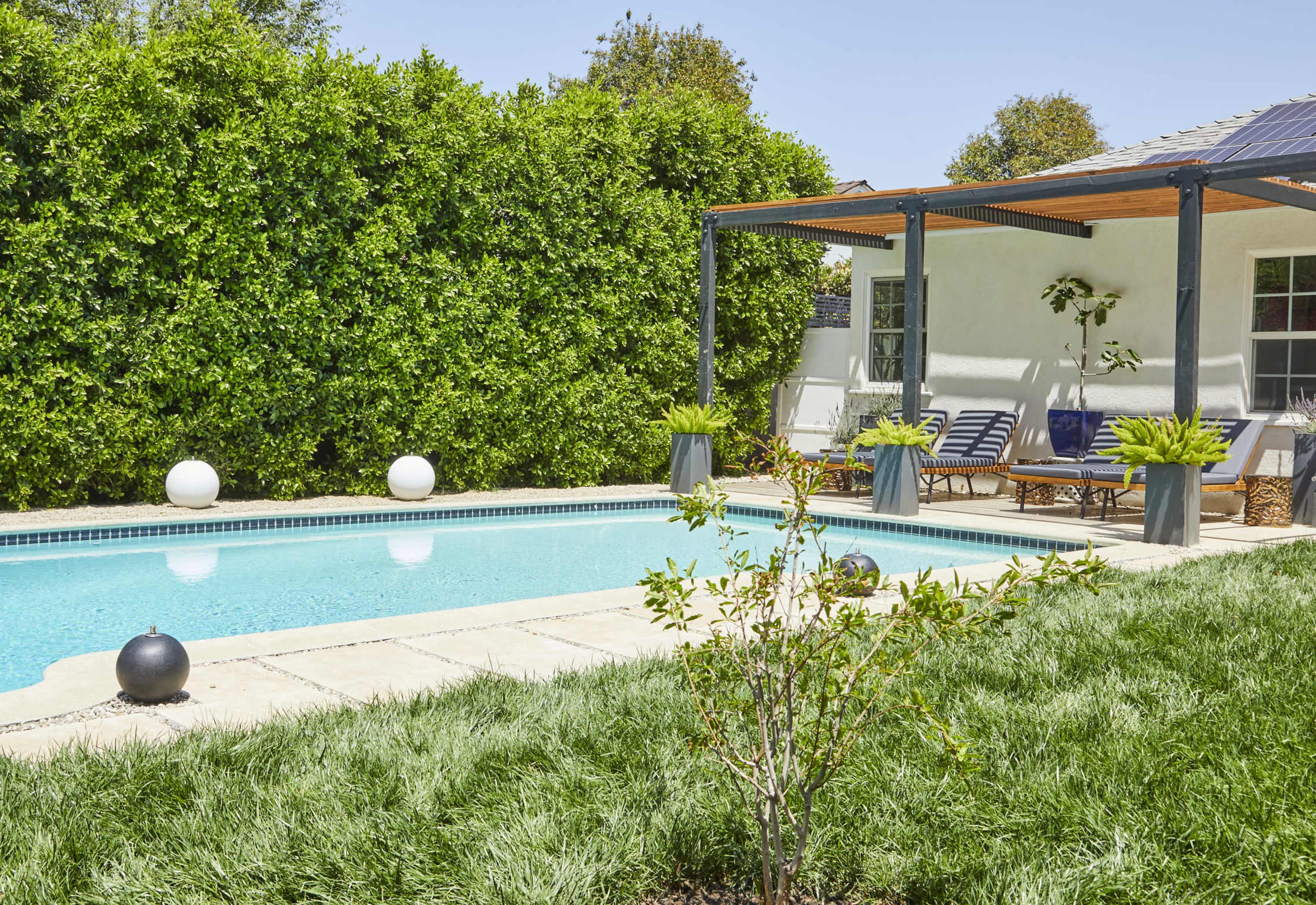 The image shows a backyard pool area surrounded by greenery with lounge chairs and a covered seating space.