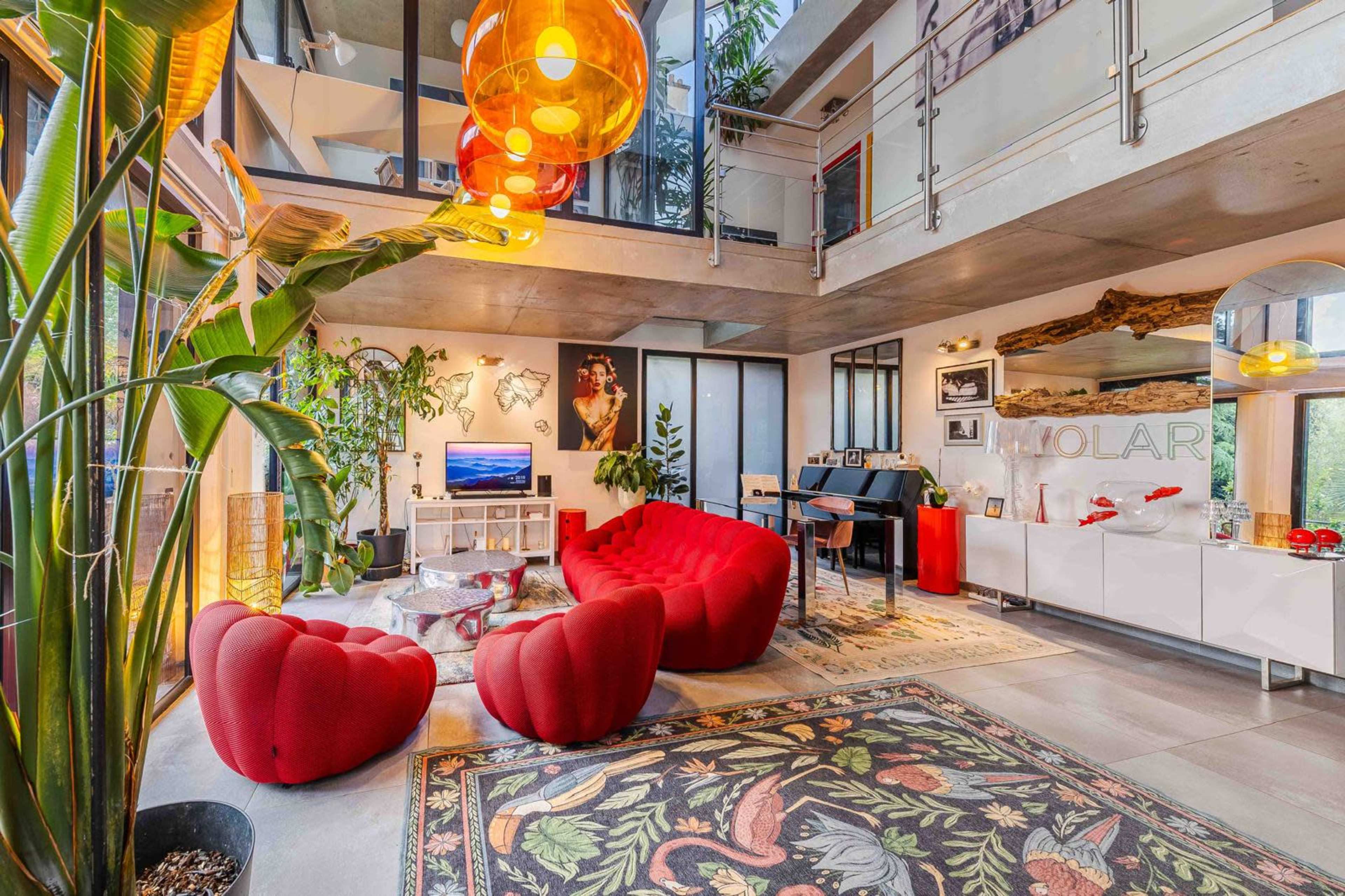 The image shows a modern, open-concept living space featuring a vibrant red sofa, a large area rug with floral patterns, and various plants, along with a minimalist kitchen area in the background.