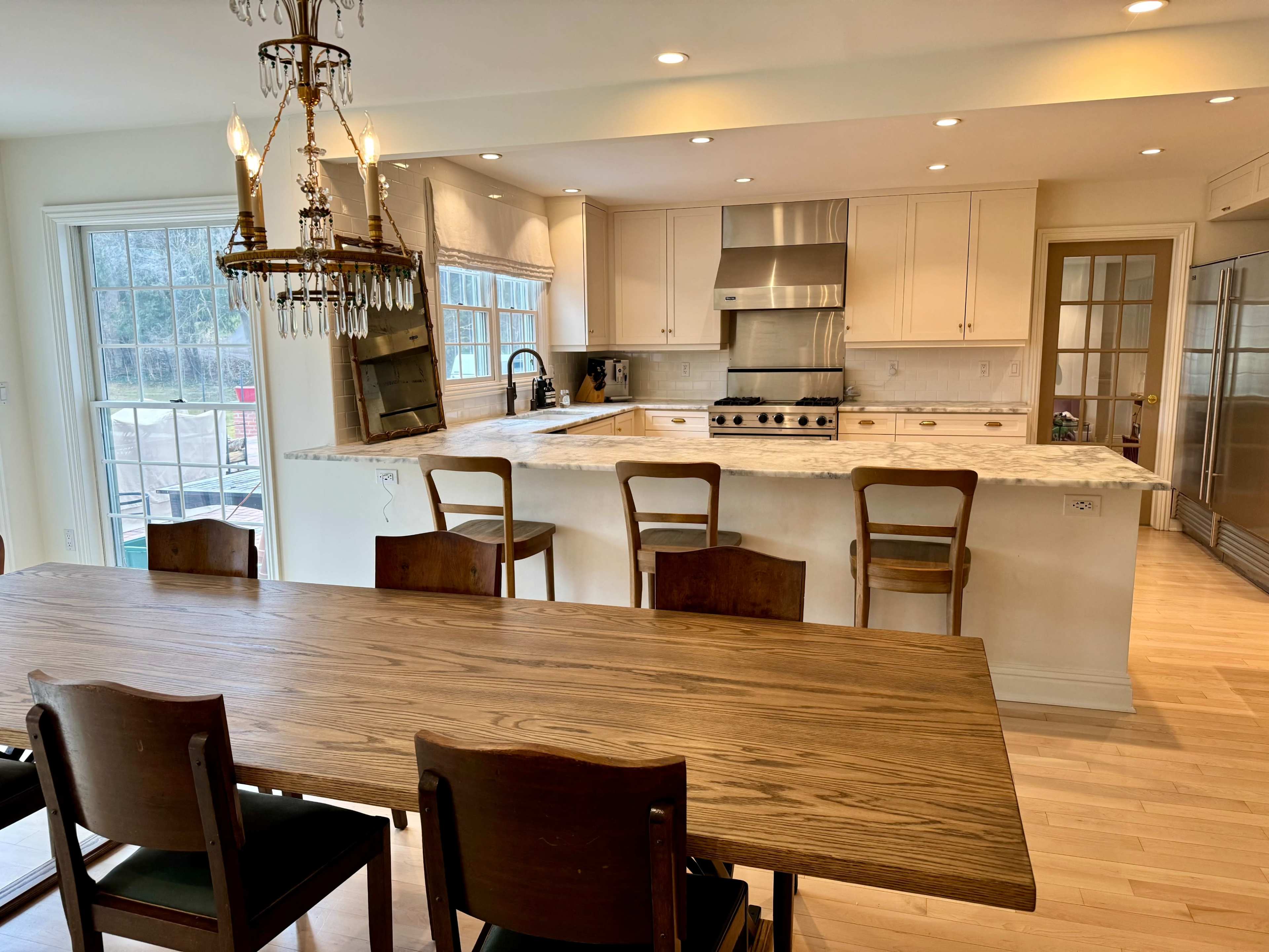 The image shows a modern kitchen with a large wooden dining table, a countertop with a stainless steel stove and range hood, and multiple windows allowing natural light to enter.