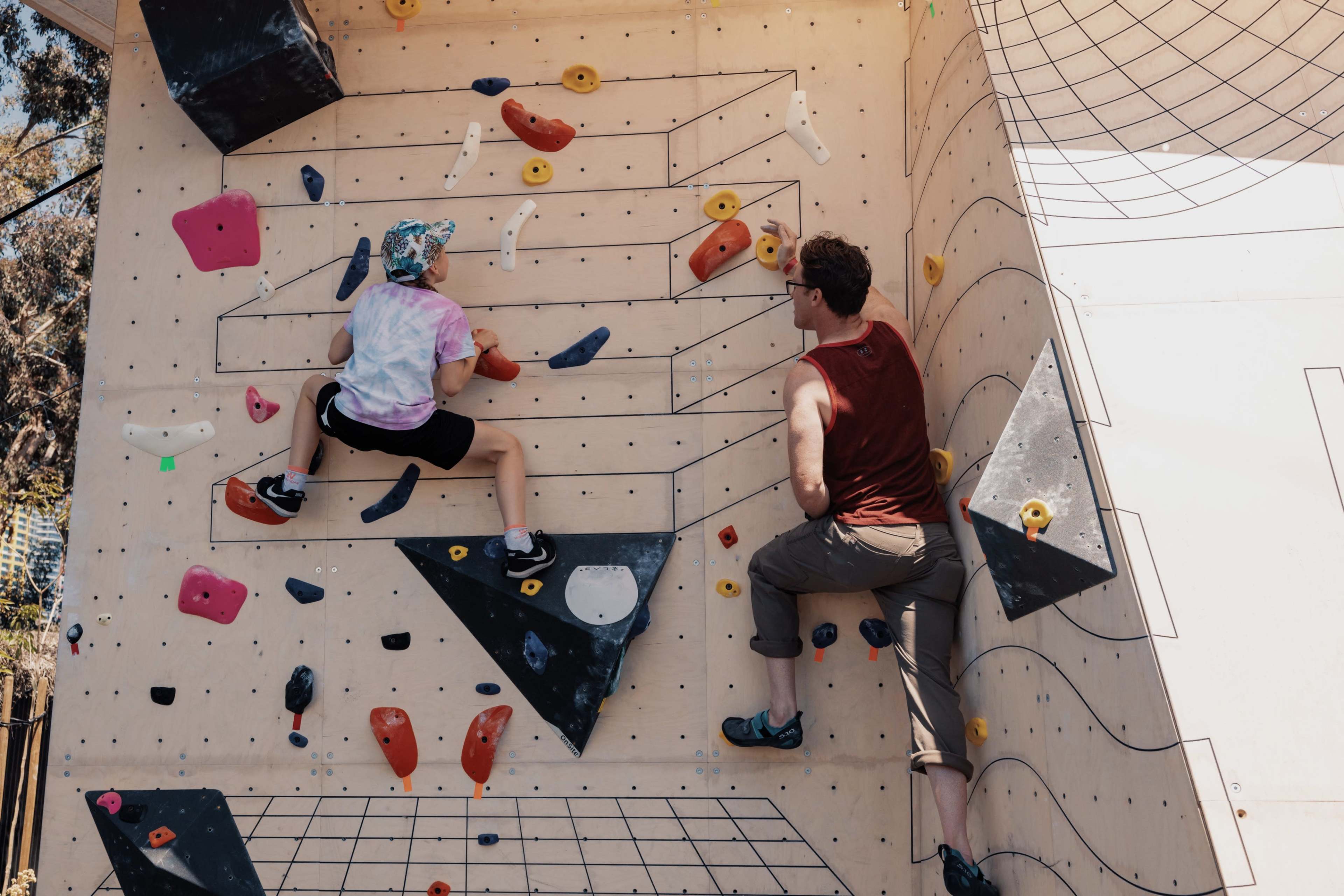 Two climbers are scaling a wooden climbing wall with various colored holds.