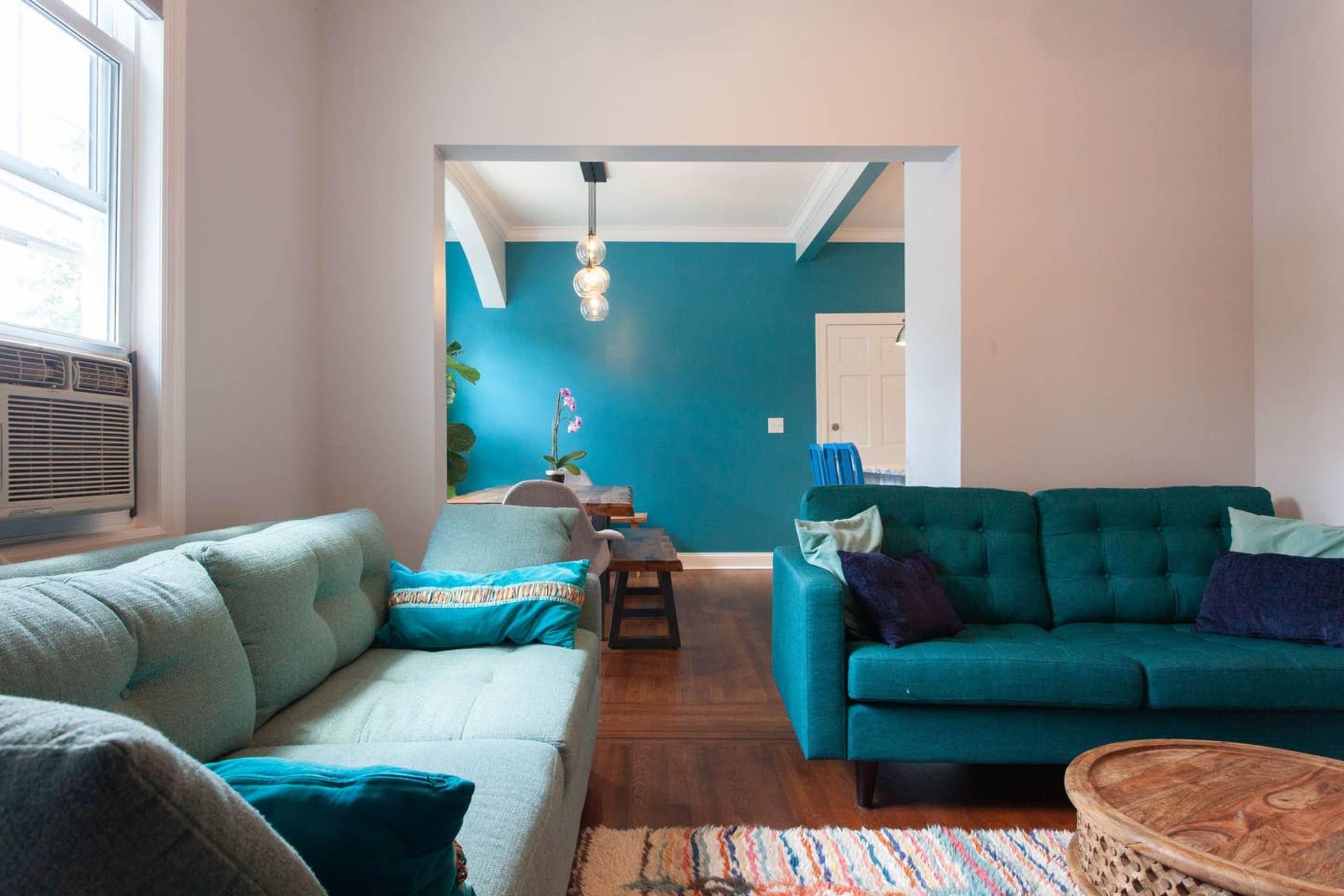 A living room features two teal sofas, a wooden coffee table, and a blue accent wall, with a doorway leading to a dining area.