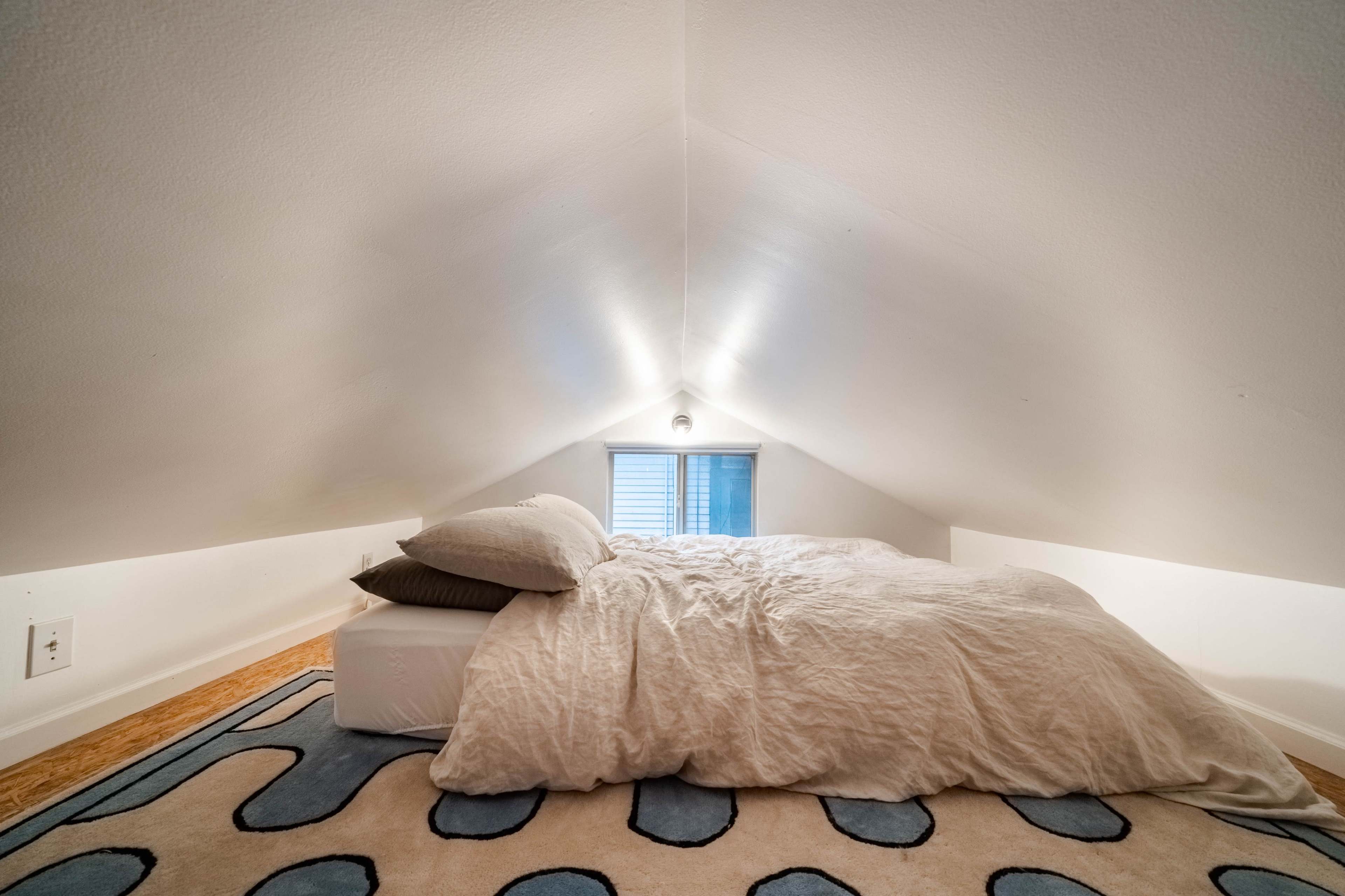 A cozy loft bedroom with a sloped ceiling, featuring a bed with a light-colored comforter and a blue patterned rug below.