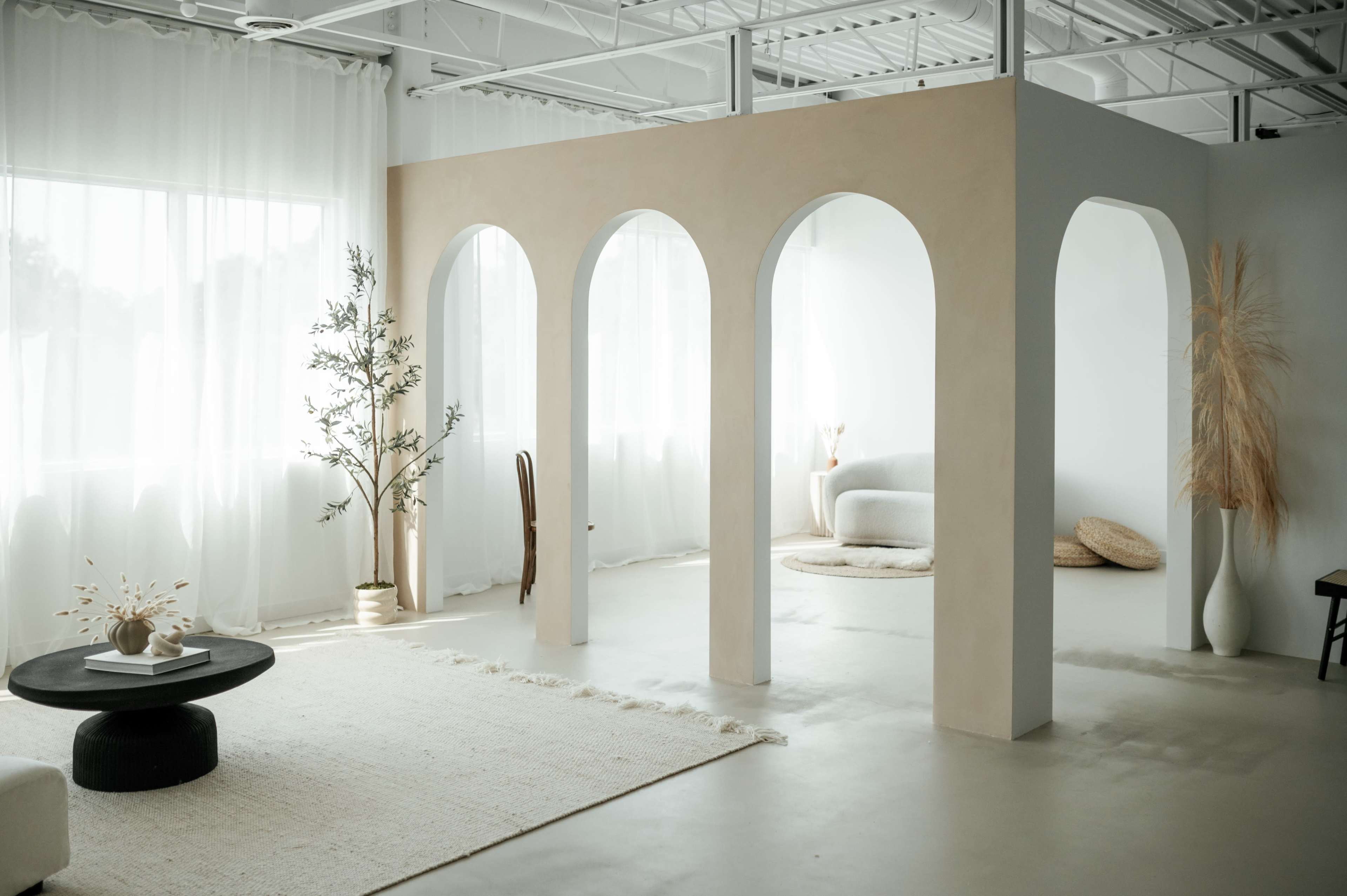 A spacious, minimalist interior features an archway structure, light-colored furnishings, and large windows allowing natural light to flood the room.