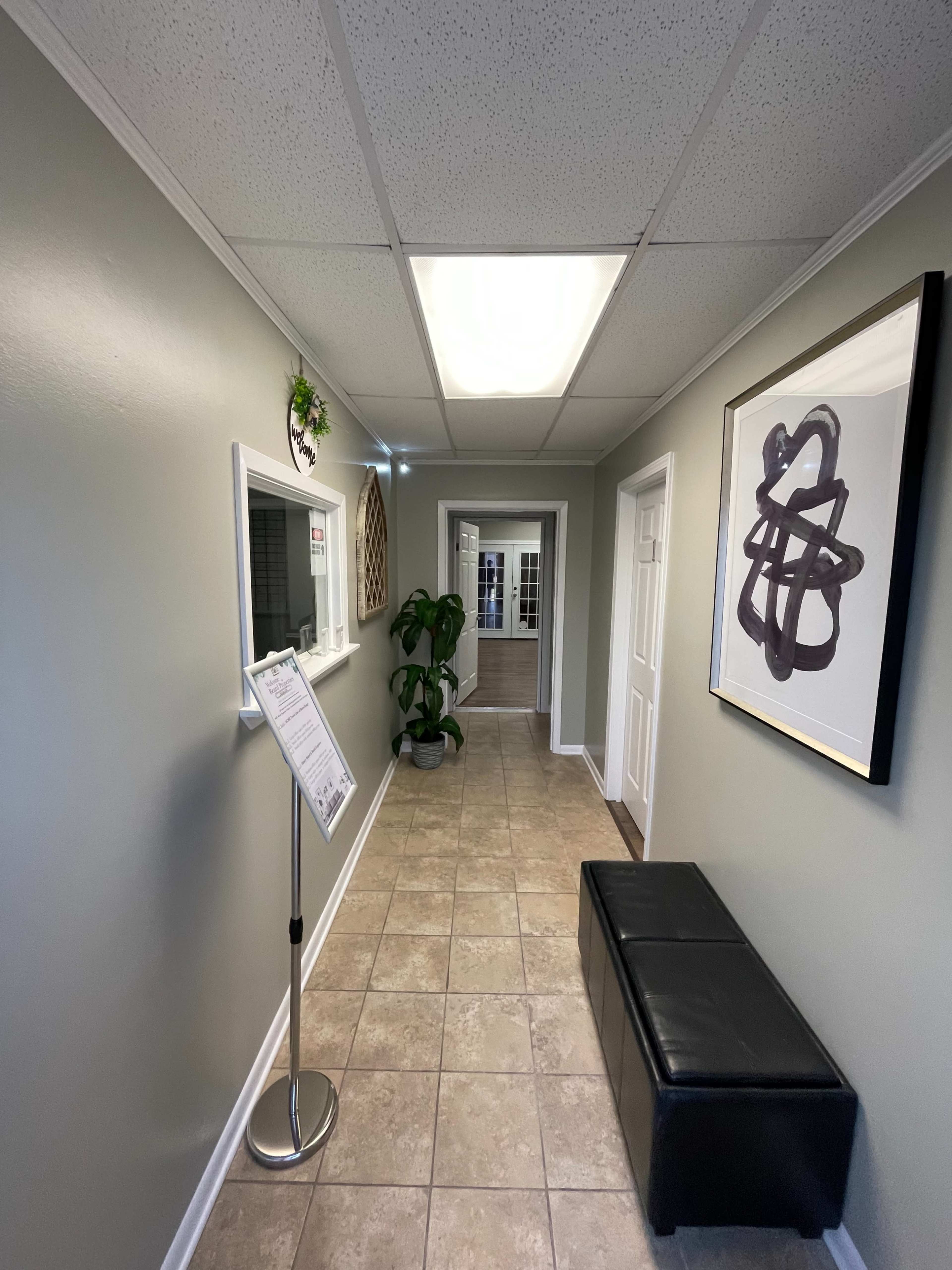 A hallway with tiled flooring, light green walls, a framed abstract artwork, a potted plant, and a menu stand near the entrance.