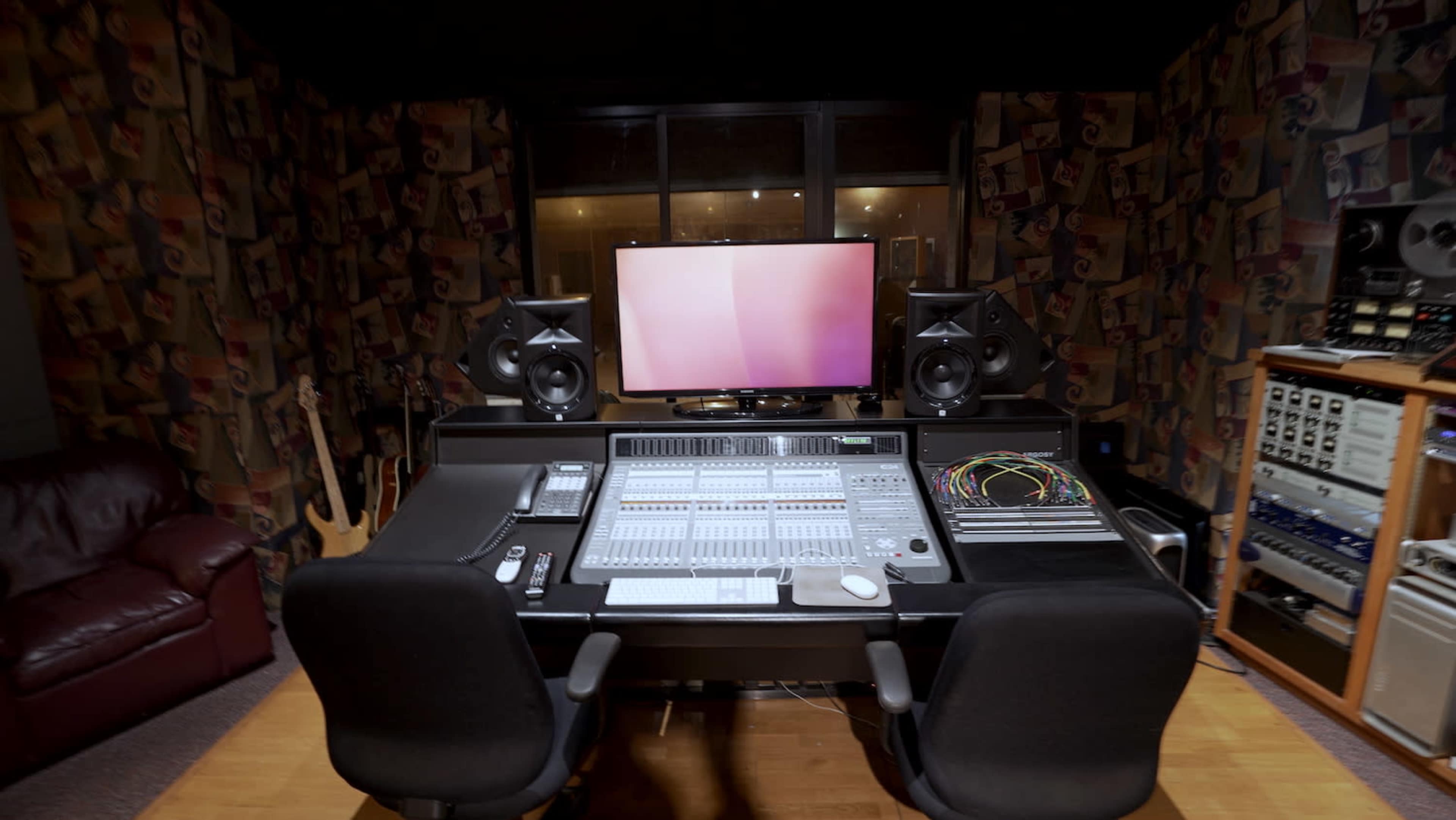 A music studio with a large mixing console, two studio monitors, a computer screen, and various audio equipment against a patterned wall.