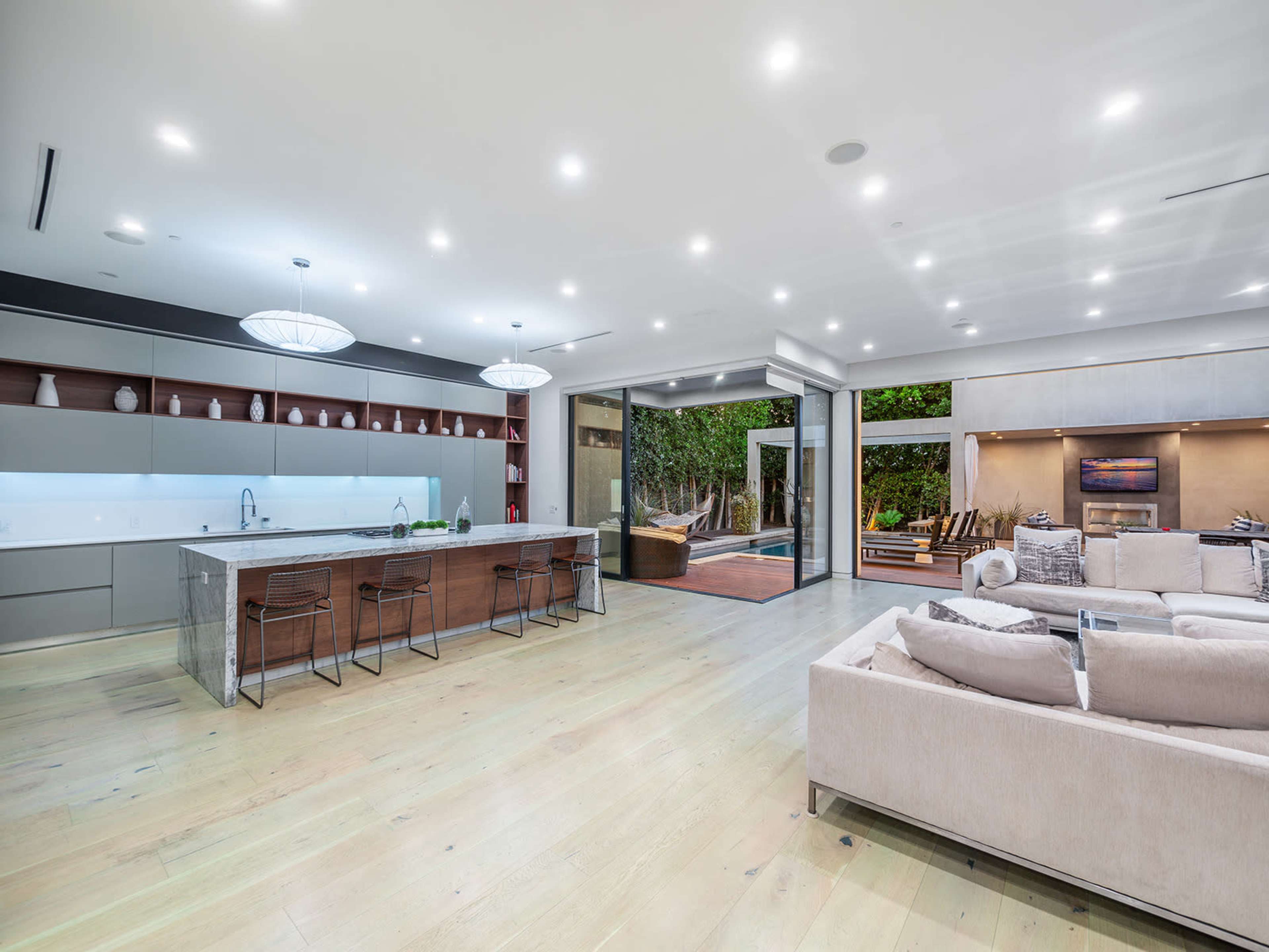 The image shows a modern open-concept living space featuring a sleek kitchen island, a living area with a large sofa, and glass doors leading to an outdoor patio.