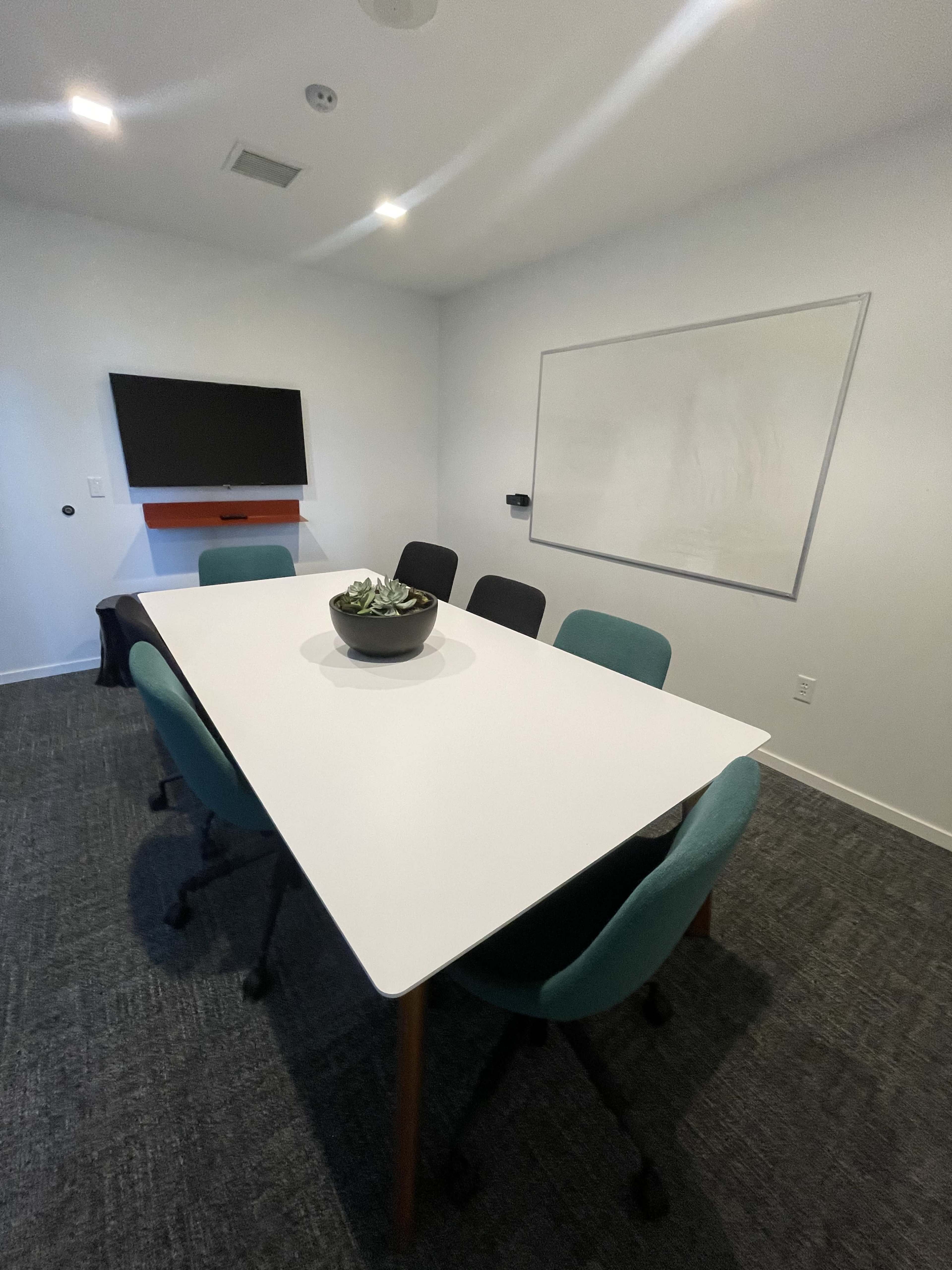 A conference room features a large white table surrounded by chairs, with a TV mounted on the wall and a blank whiteboard.