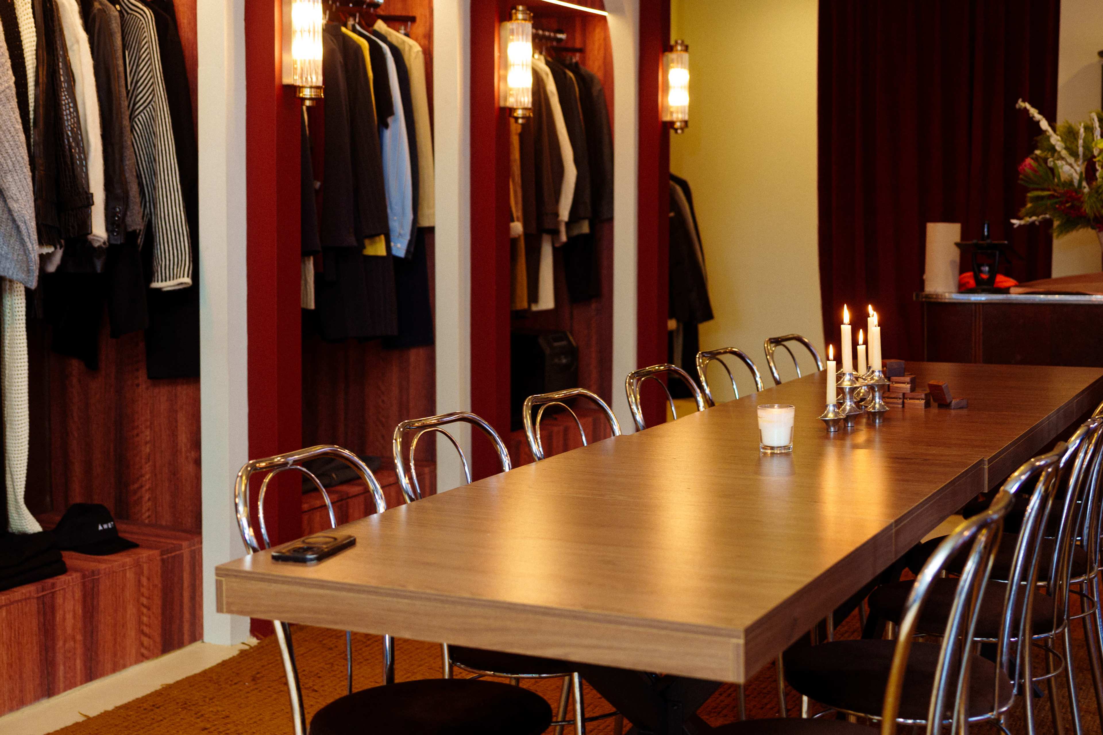 A long wooden table with candles is set in a room featuring clothing displayed on racks and shelves along the walls.