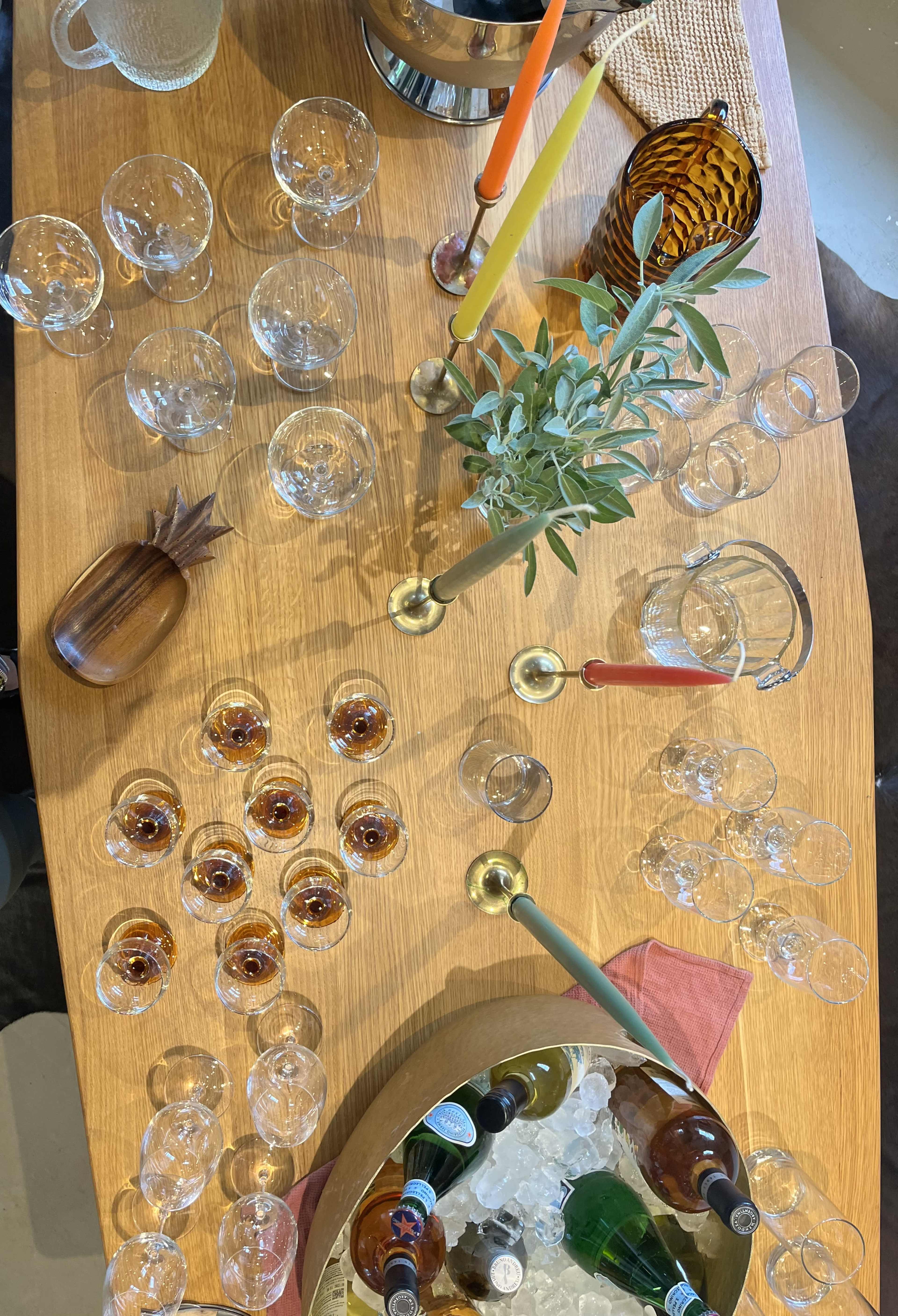 An overhead view of a wooden table set with various glasses, colorful candles, a plant centerpiece, and a bowl of ice containing assorted bottles.