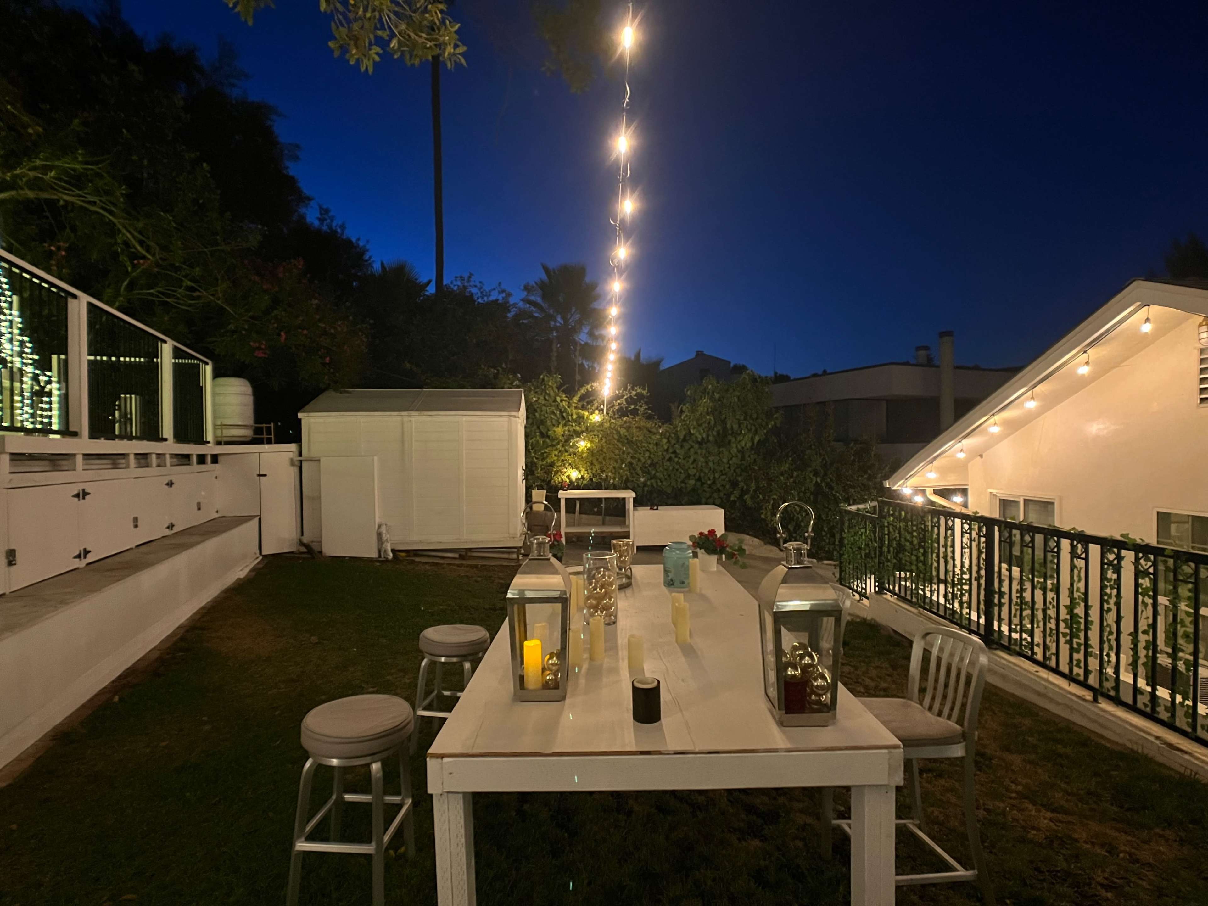 A white-table setting with candles and lanterns is arranged on a grassy area under string lights in a backyard at night.