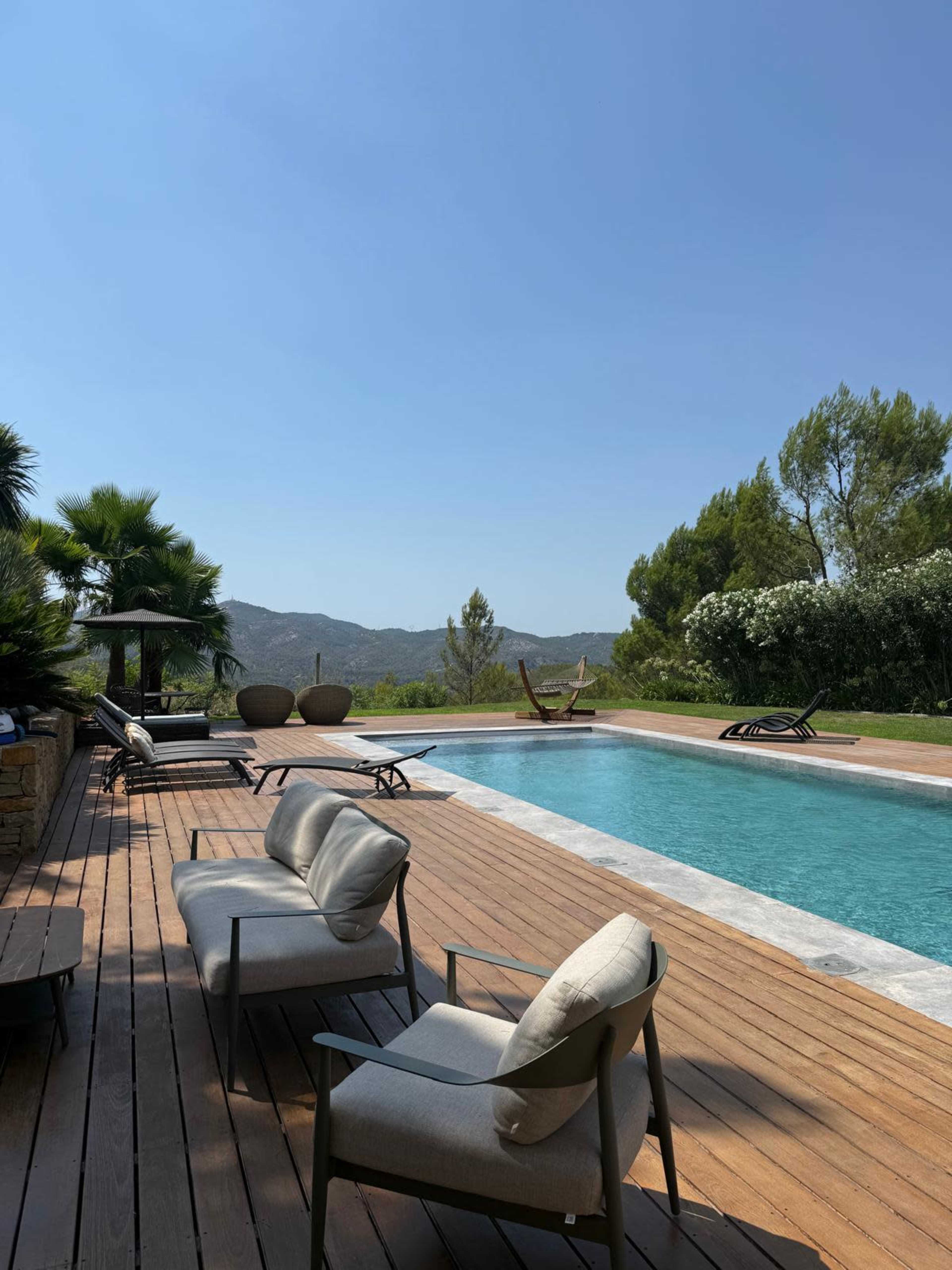 The image shows a tranquil outdoor pool area with wooden decking, lounge chairs, and surrounding greenery under a clear blue sky.