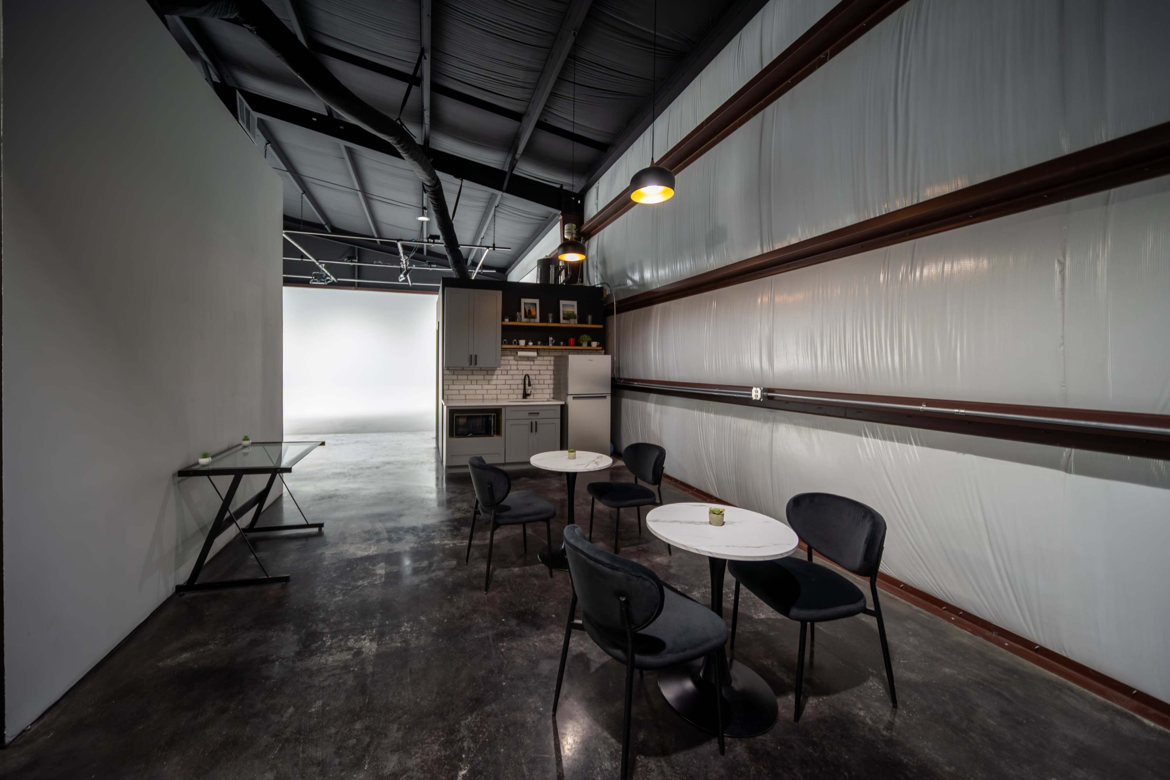 The image shows a modern, industrial-style interior featuring a small seating area with black chairs and tables, a kitchenette, and white walls that connect to a spacious lighting area.