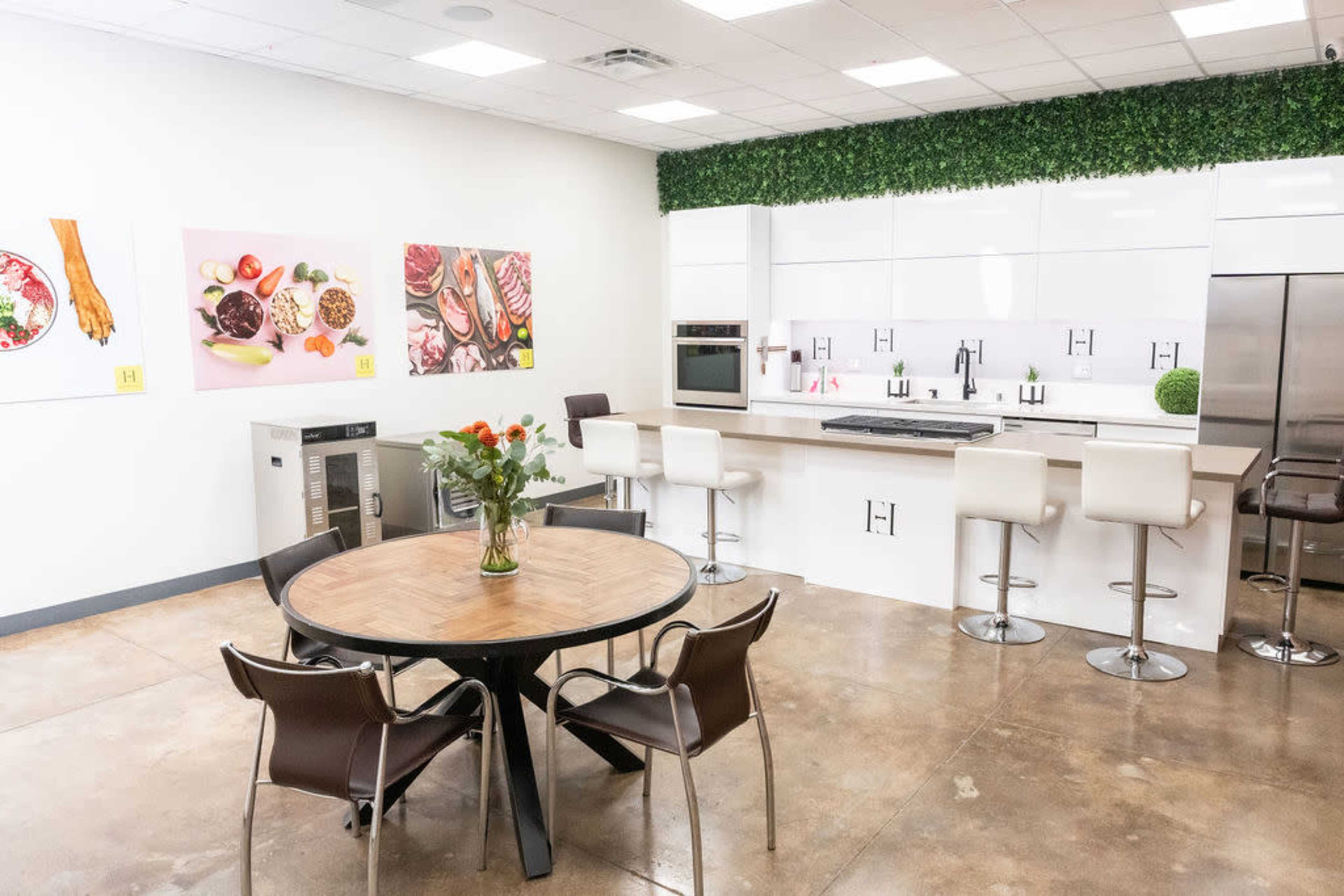 The image shows a modern kitchen space with a round table and chairs, featuring a cooking area with sleek white cabinetry and greenery decor along the walls.