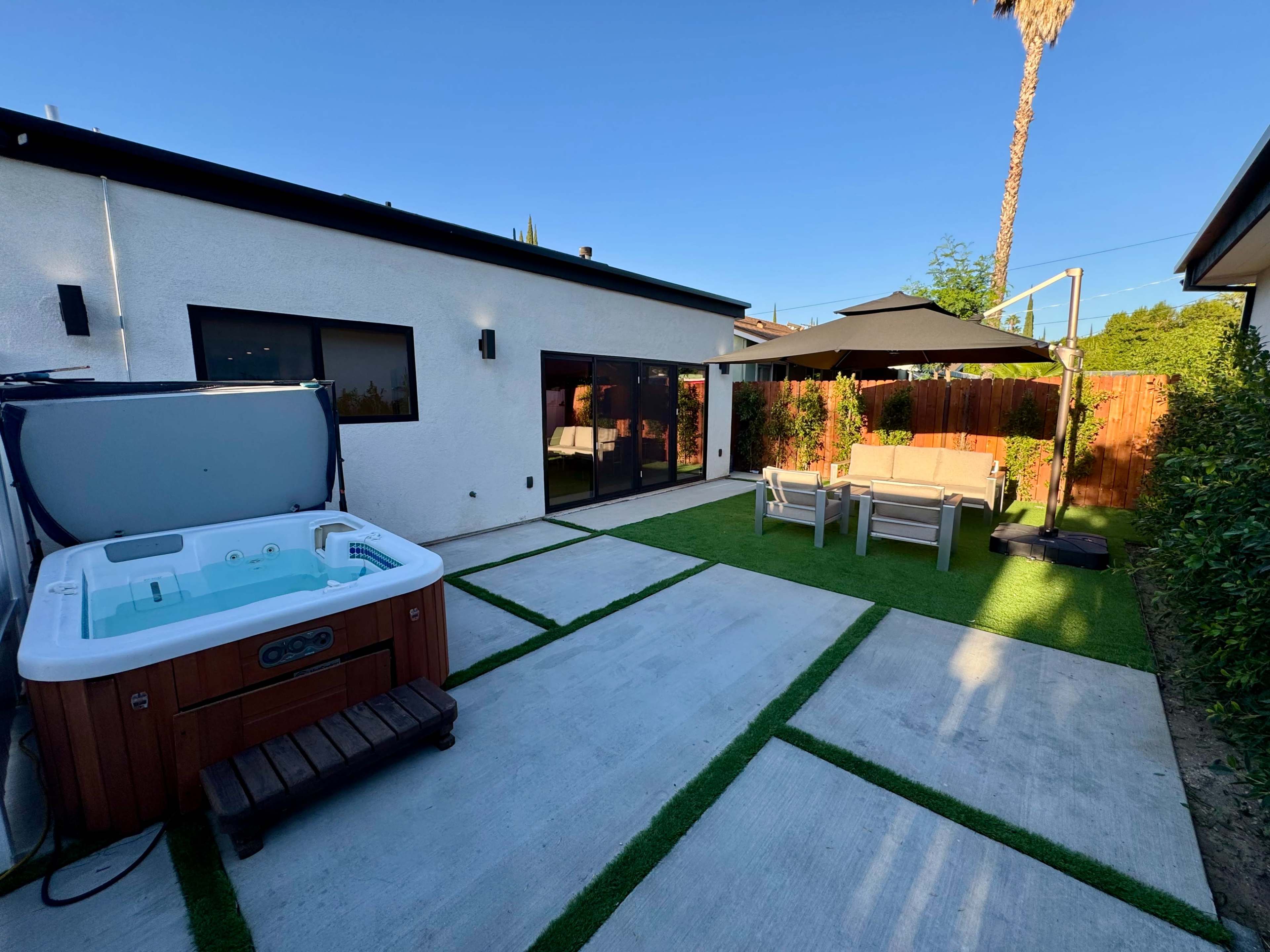 The image shows a backyard with a hot tub, a shaded seating area, and concrete pathways bordered by grass.