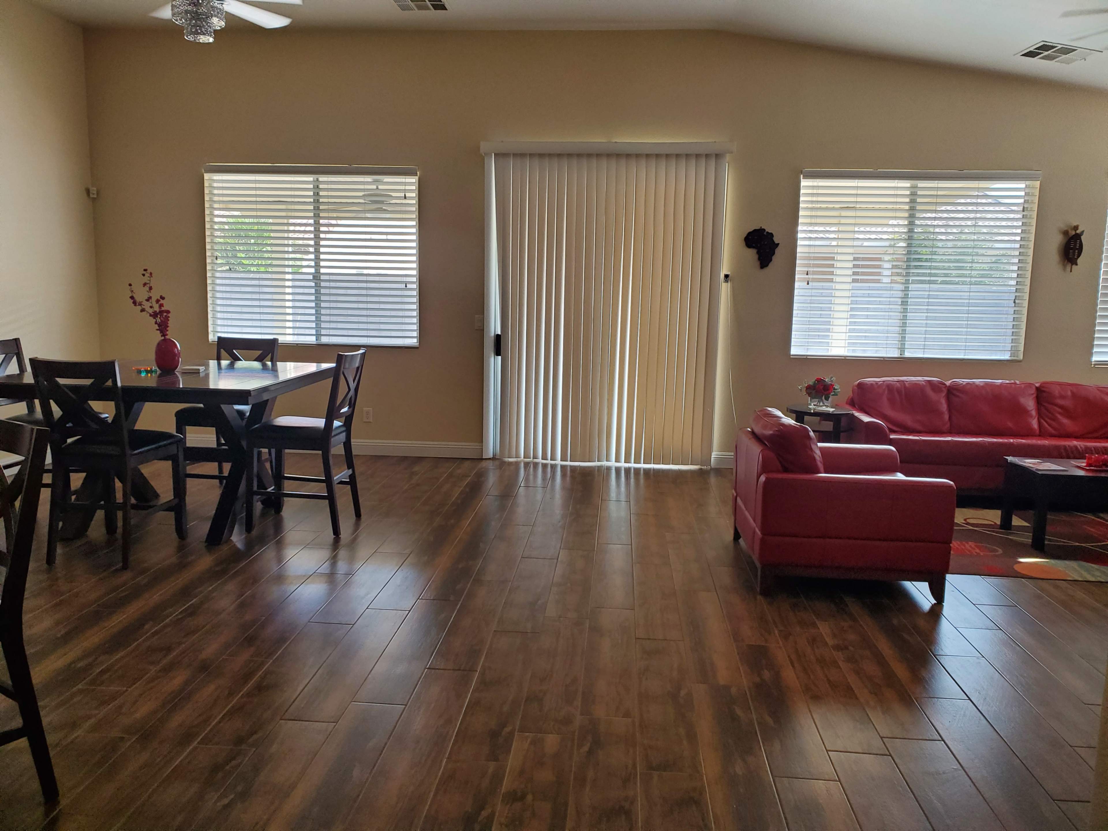 A spacious living area with a dining table and chairs on one side and a red sofa on the other, featuring large windows with blinds.