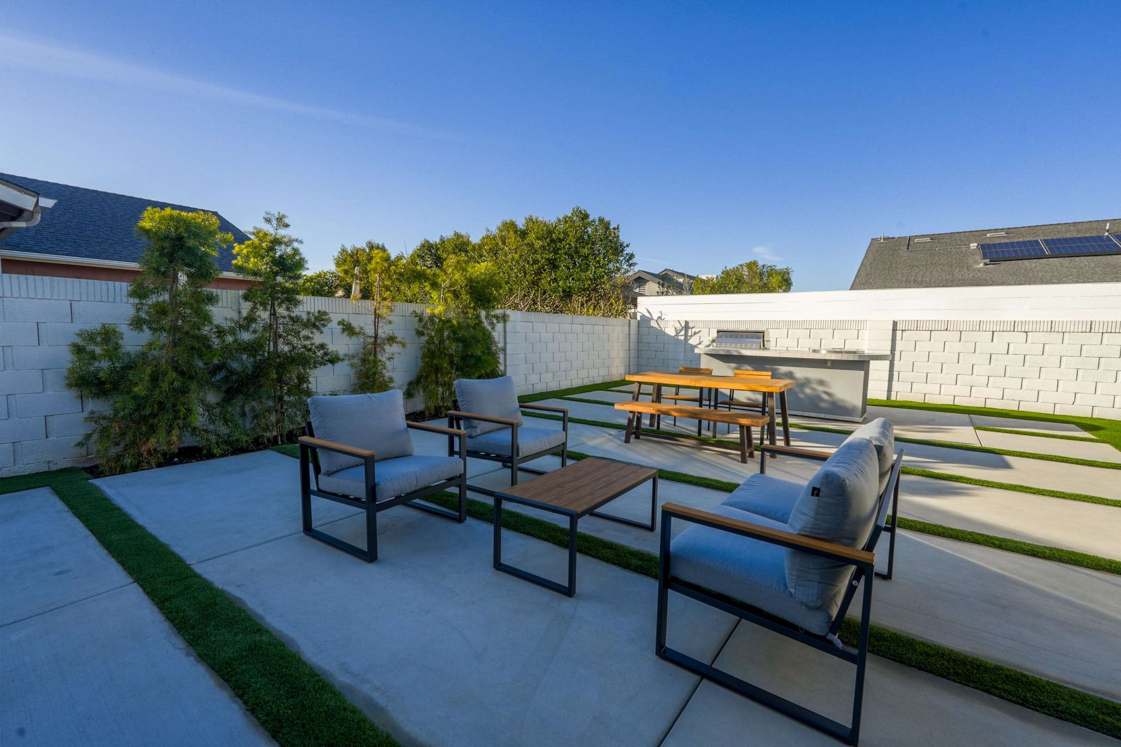 A modern outdoor patio features four metal-framed chairs with cushions and a wooden table set on a concrete surface surrounded by artificial grass and a landscaped privacy wall.