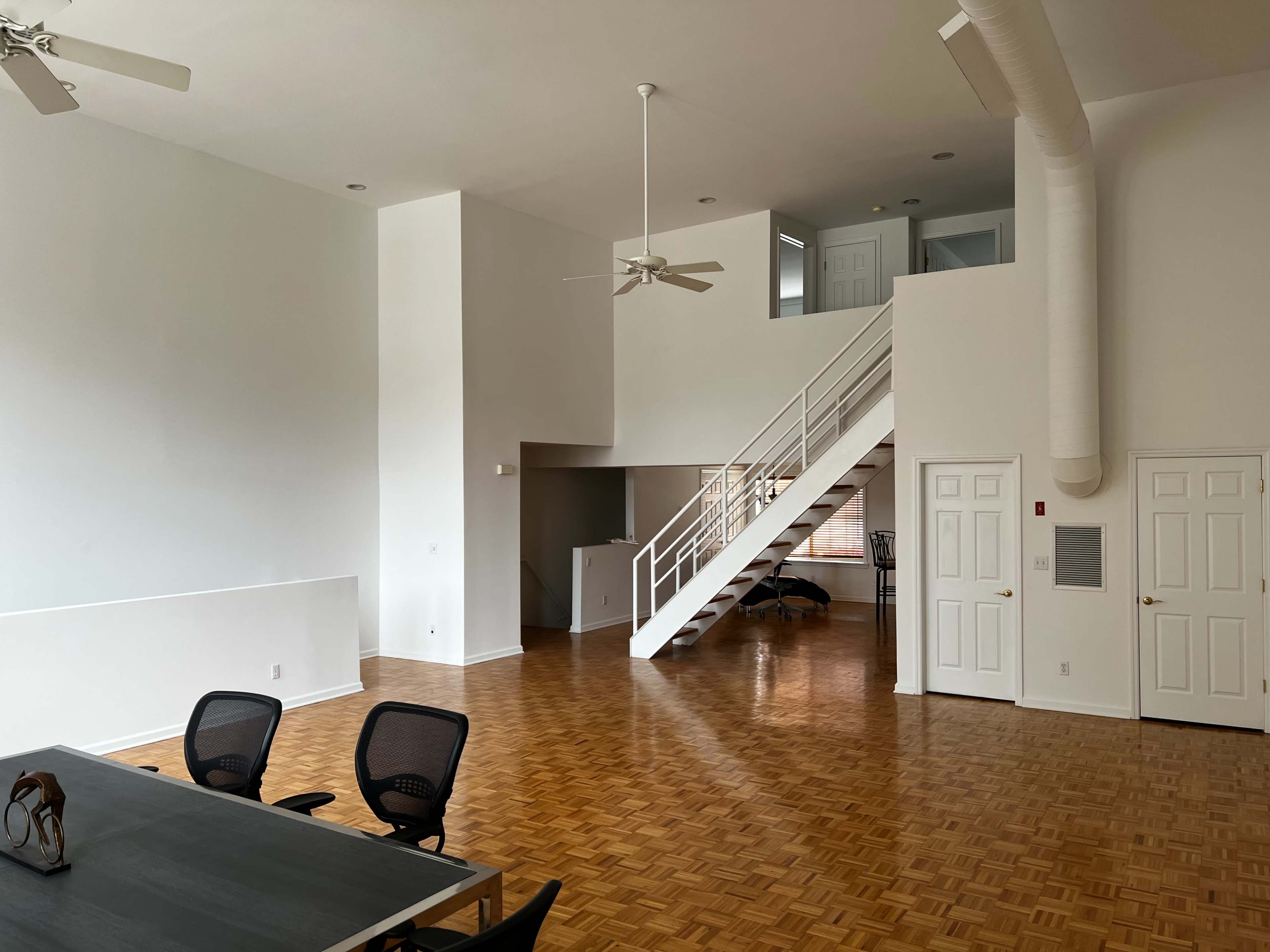 A spacious, open loft-style room with wooden flooring, a staircase leading to a second level, and a large window.
