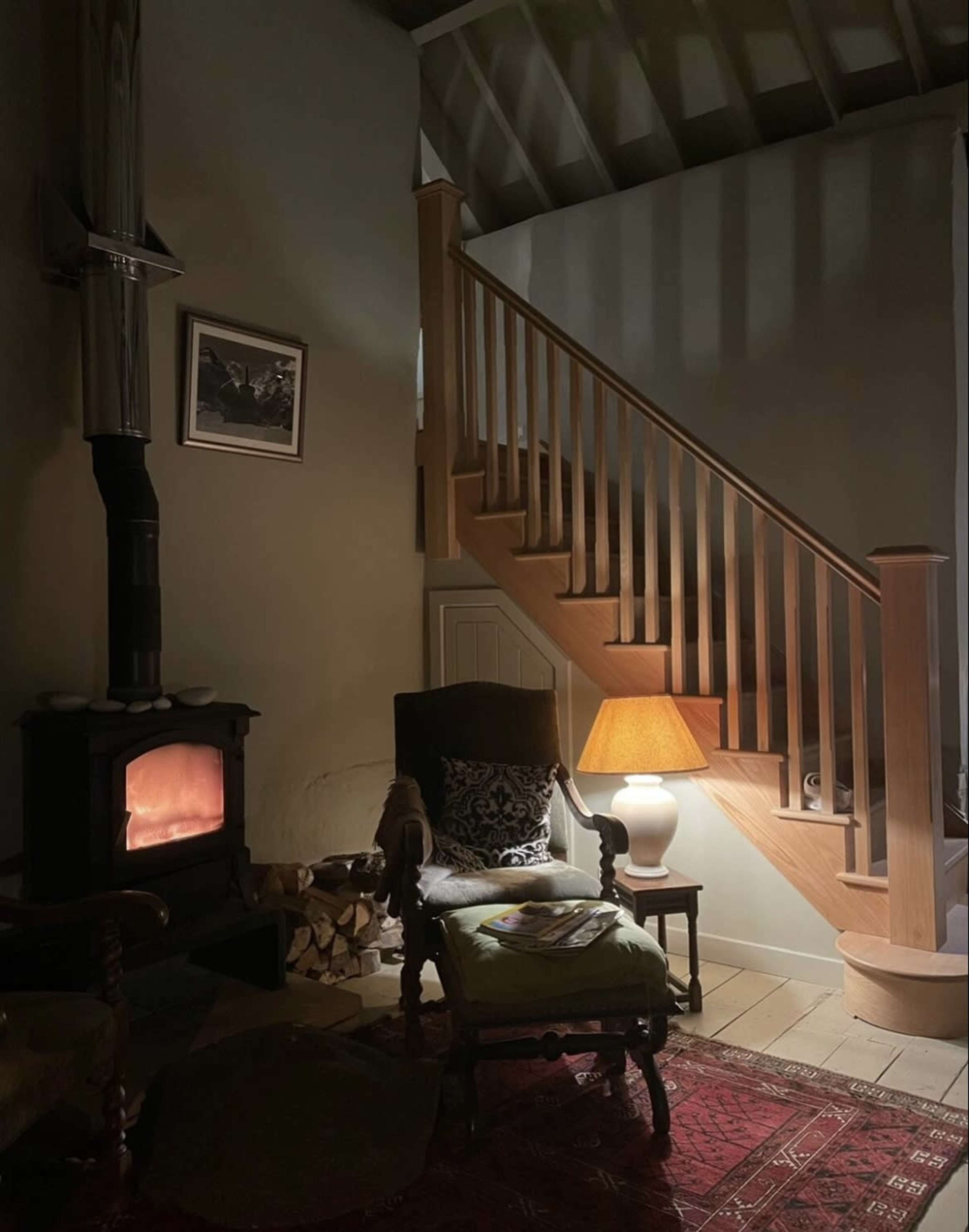 A cozy interior features a wooden staircase, an armchair beside a lamp, and a stove with glowing embers.
