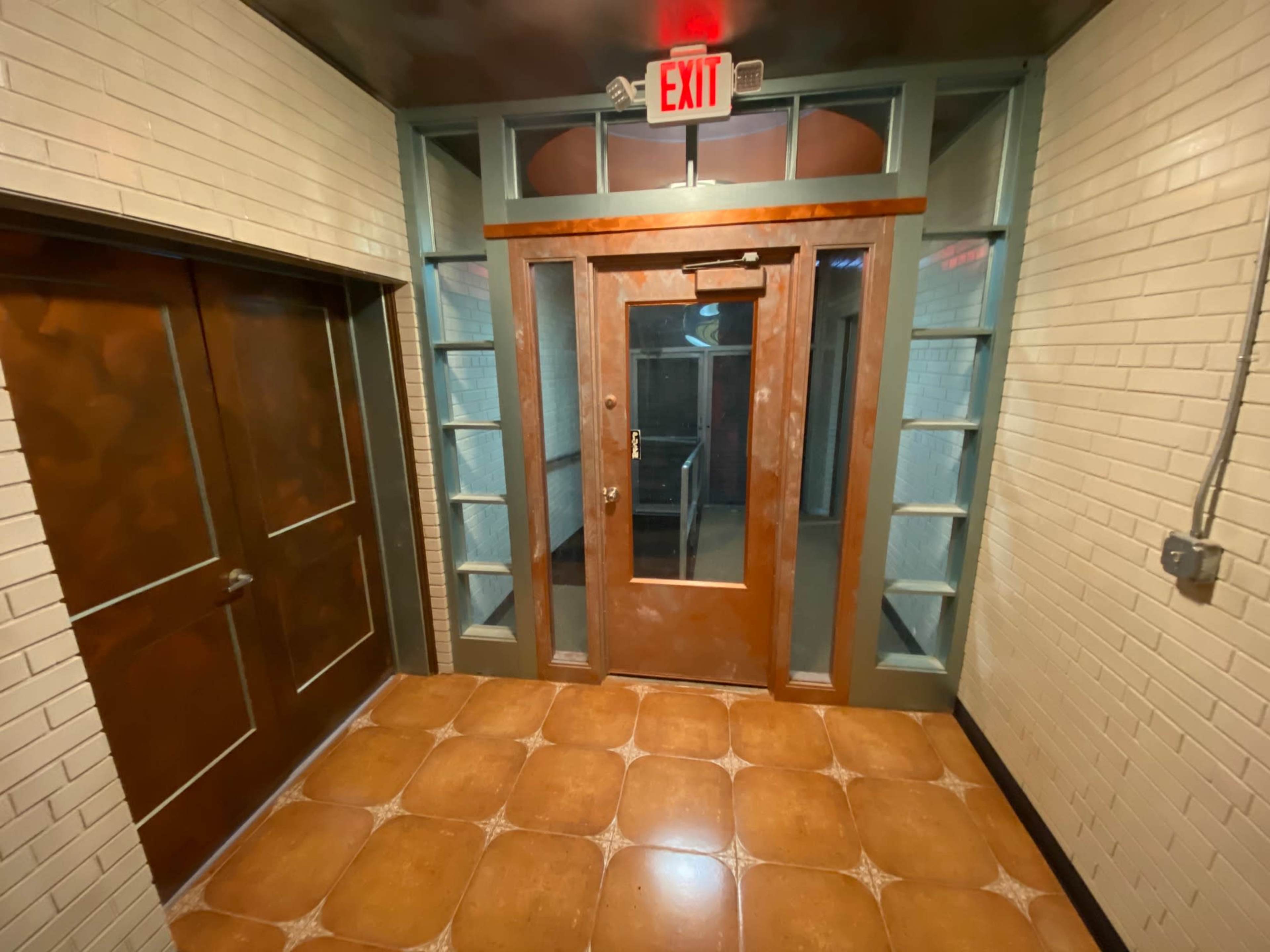 A hallway with a wooden door, tiles on the floor, and an exit sign above the door.