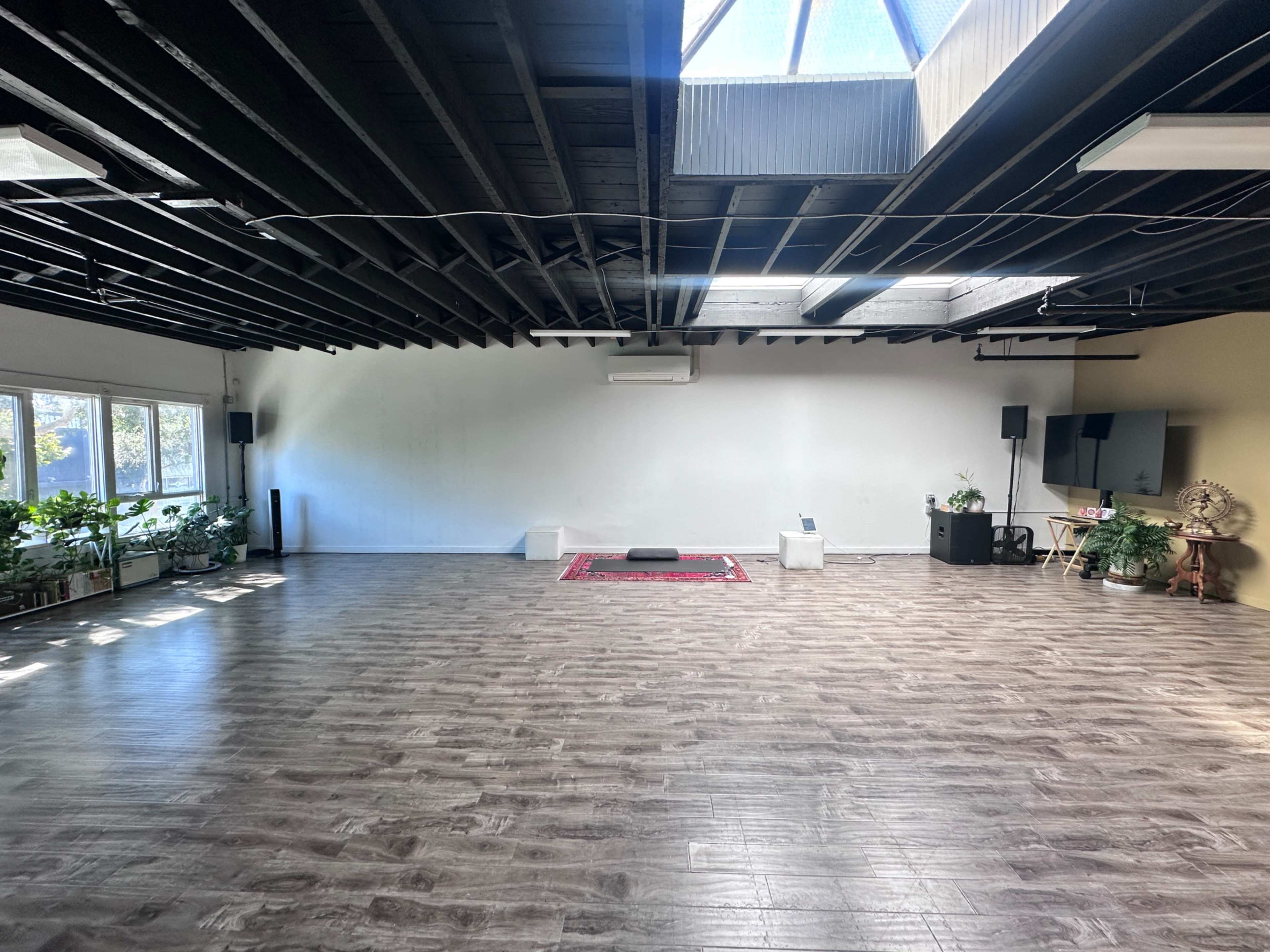 The image shows a spacious, well-lit room with wooden flooring, large windows filled with greenery, and minimal furnishings, including a red carpet and speakers.