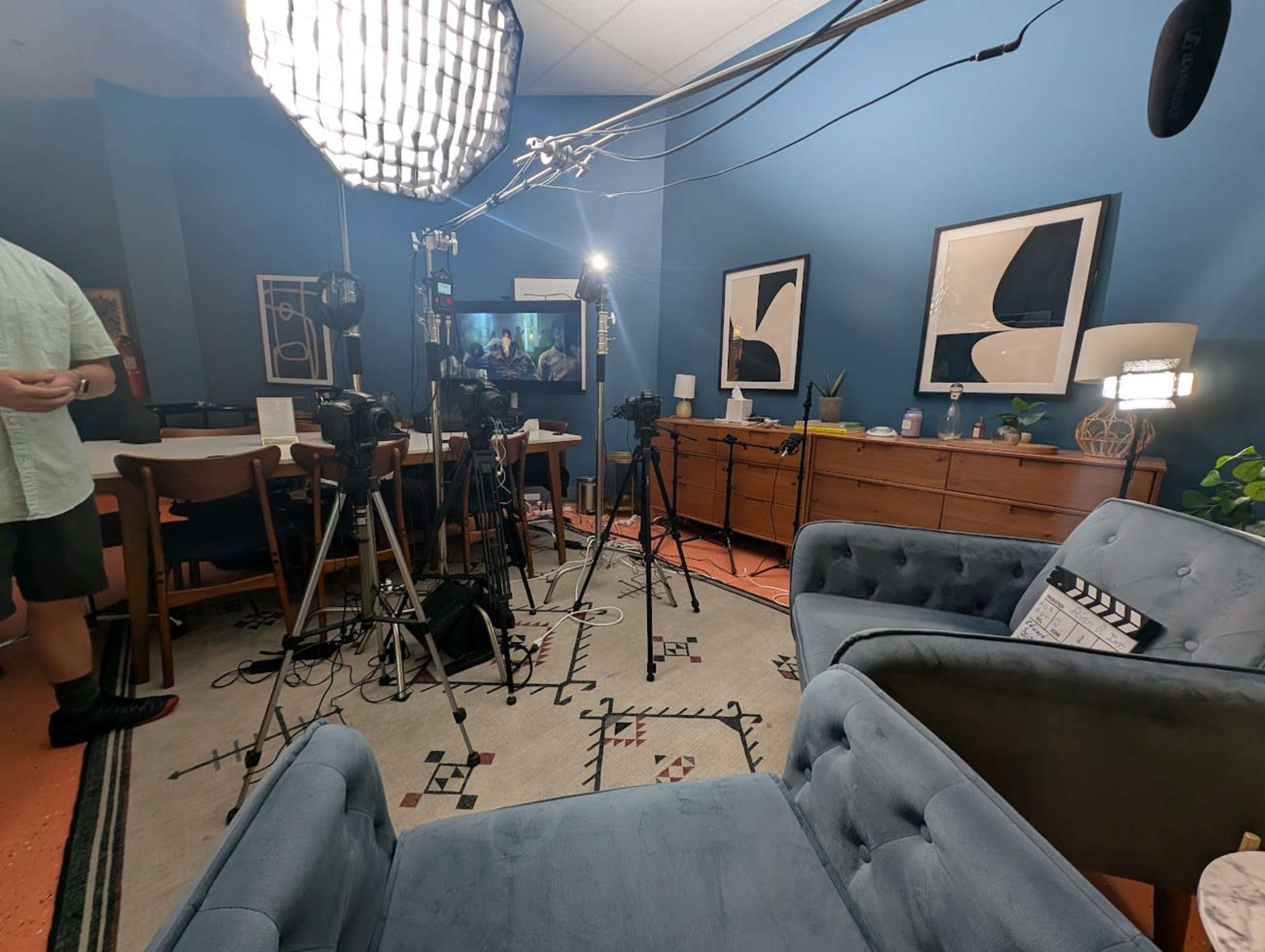 The image shows a well-lit room set up for a video production, featuring cameras and lighting equipment around a pair of blue sofas and a decorated wall.