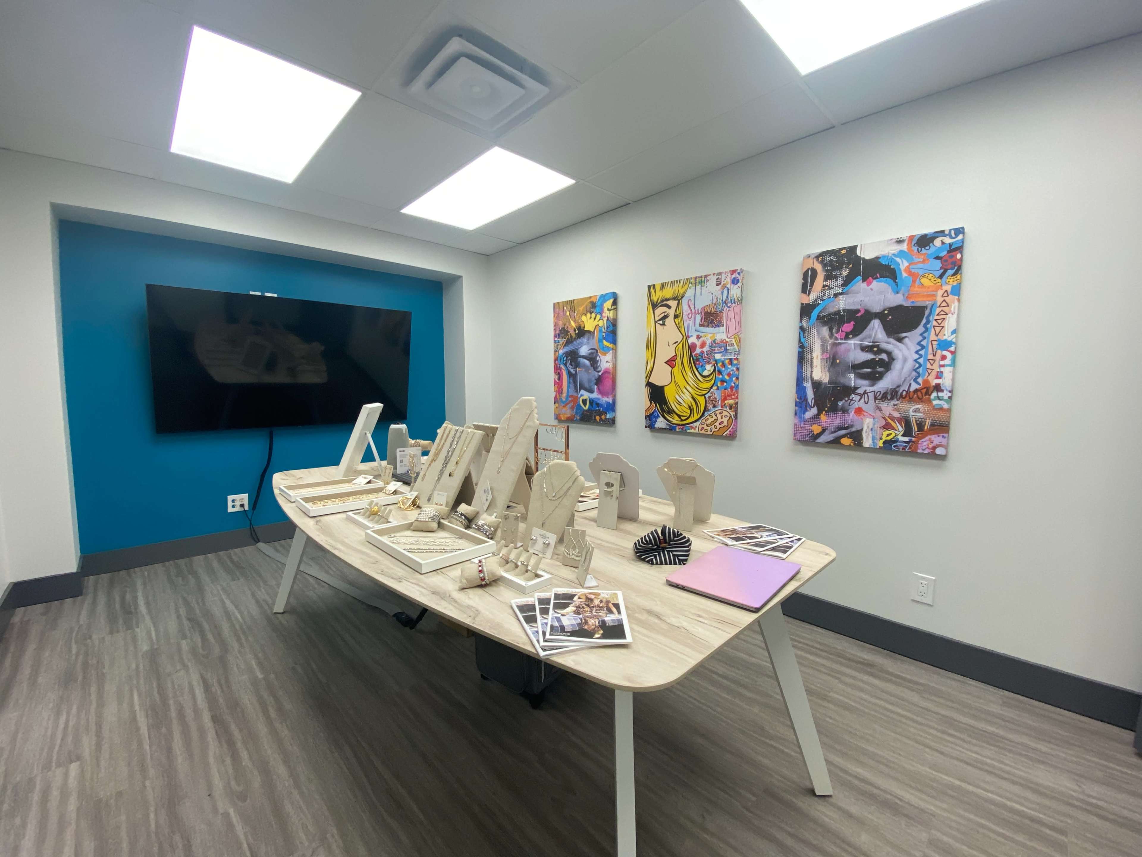 A modern conference room features a large table displaying jewelry and art, surrounded by colorful wall art and a television.