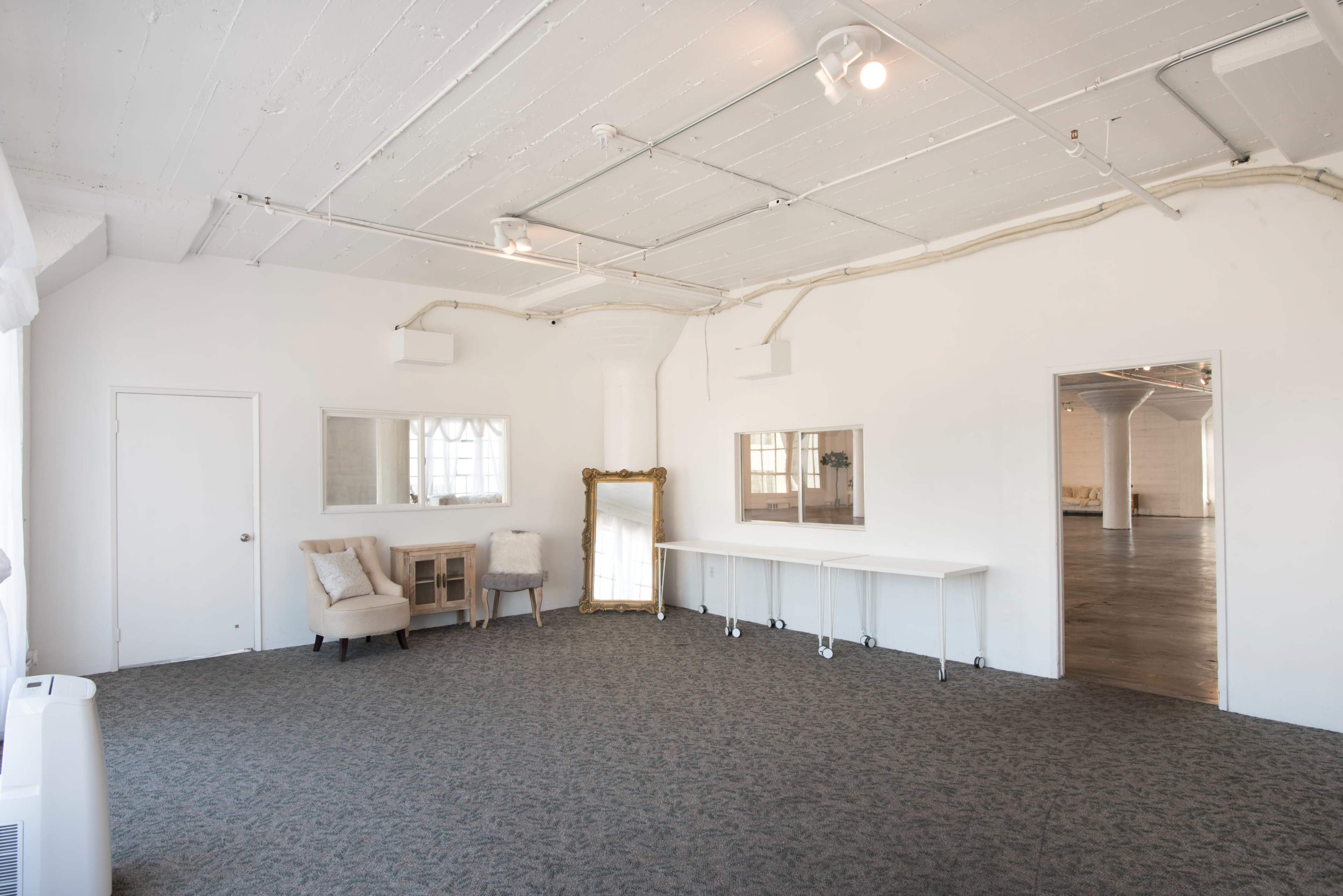 A spacious, bright room with white walls, a carpeted floor, and large mirrors along one side, featuring simple furniture including two chairs and a table.