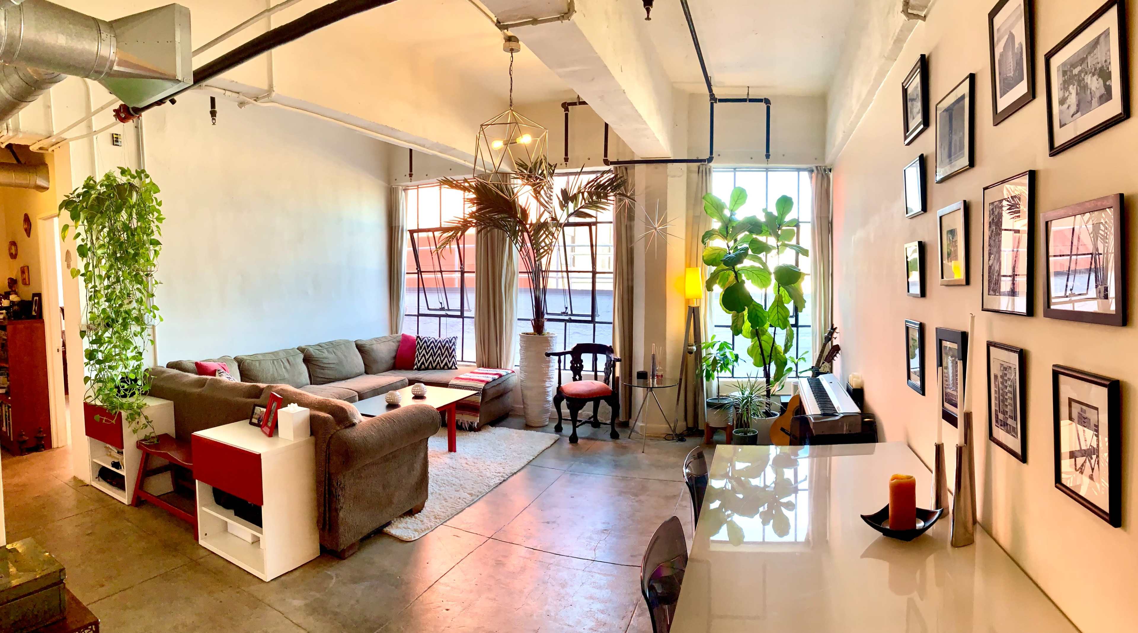 The image shows a spacious living area with a sofa, a dining table, plants, and a wall adorned with black and white framed photographs.