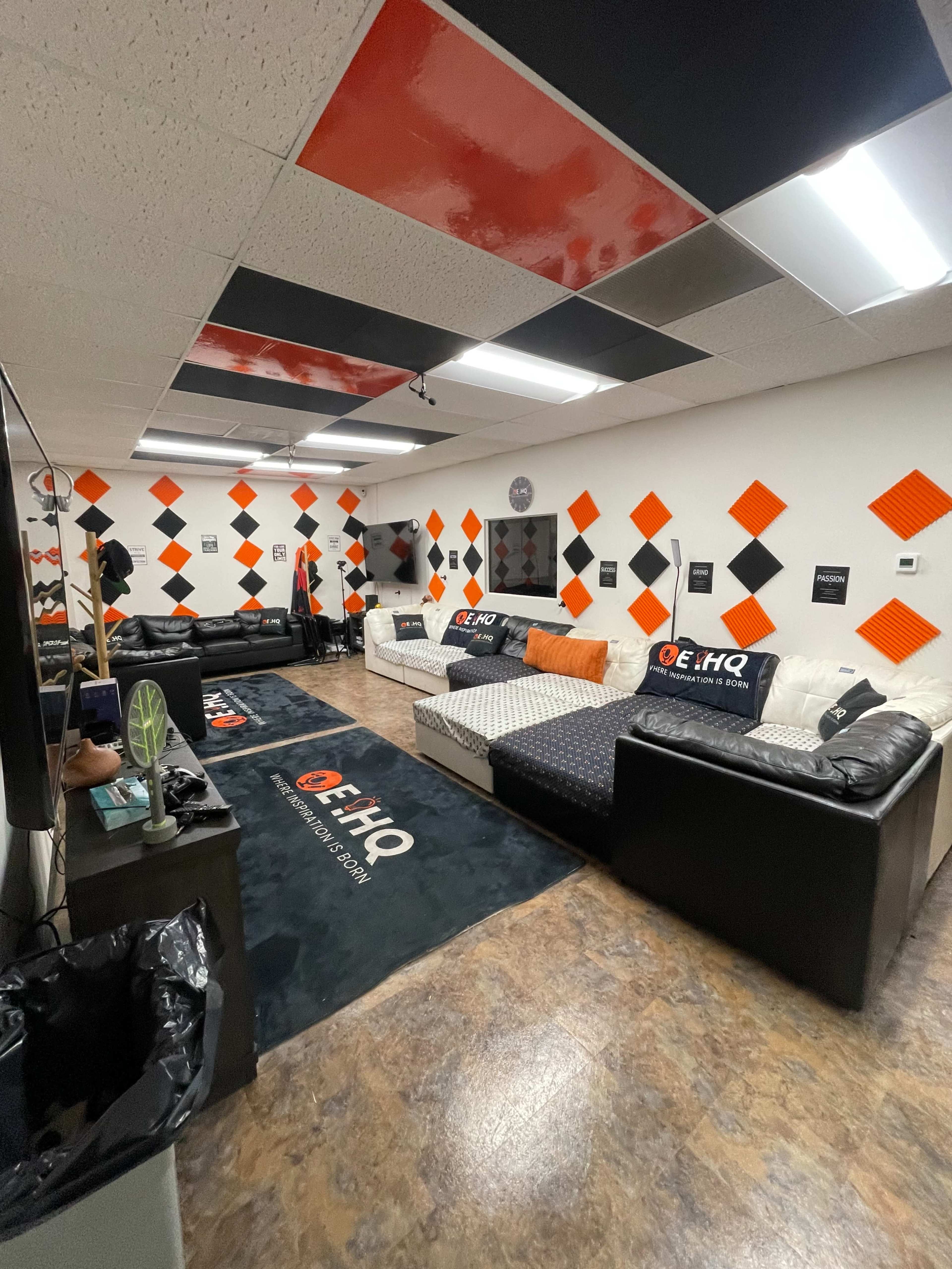 The image shows a lounge area with black and orange decor, featuring two couches, walls adorned with diamond-shaped designs, and a carpet branded with "E HQ."