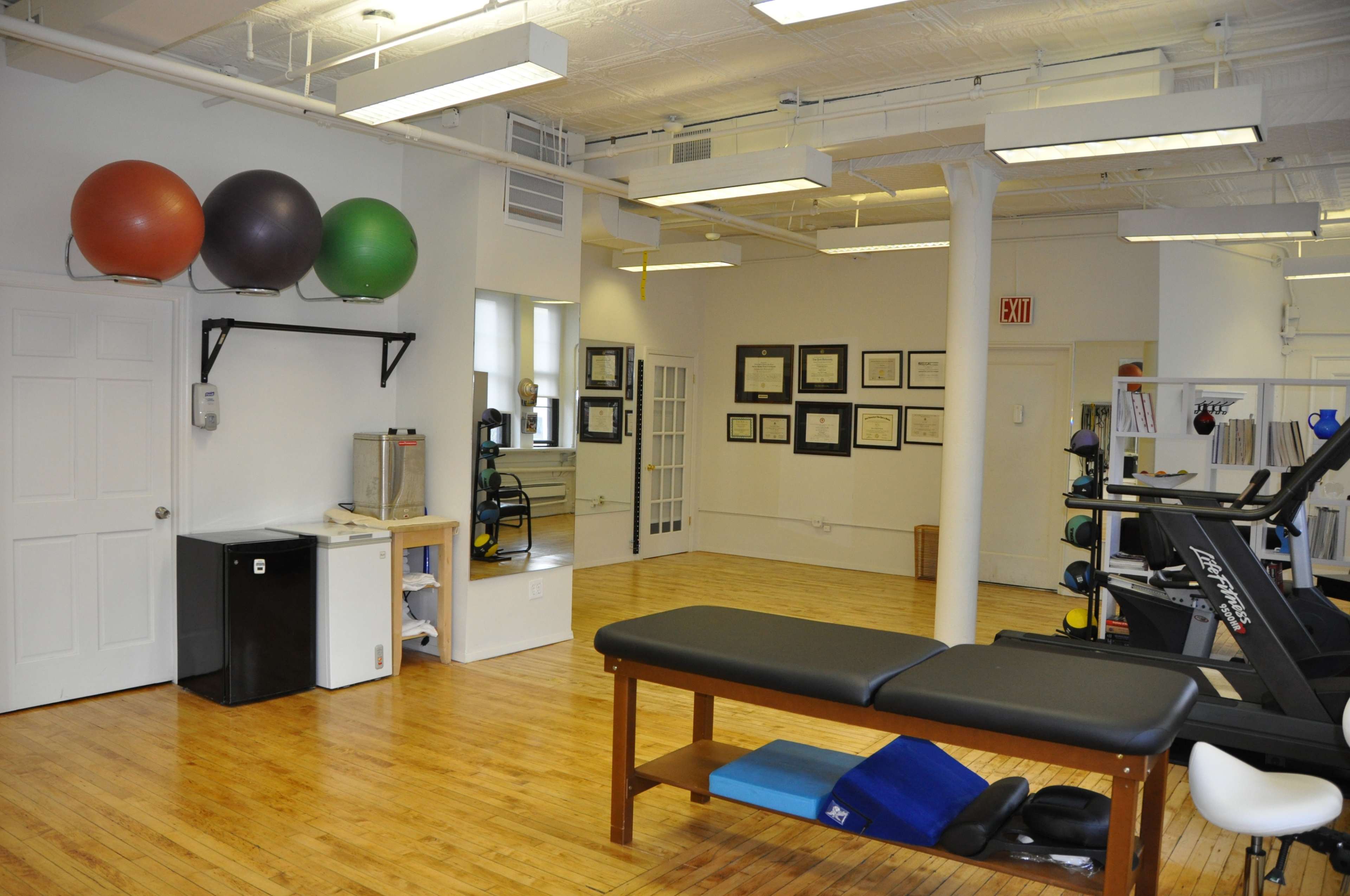 A spacious fitness studio with exercise balls mounted on the wall, a treatment table, and various workout equipment arranged in a well-lit room with wooden flooring.