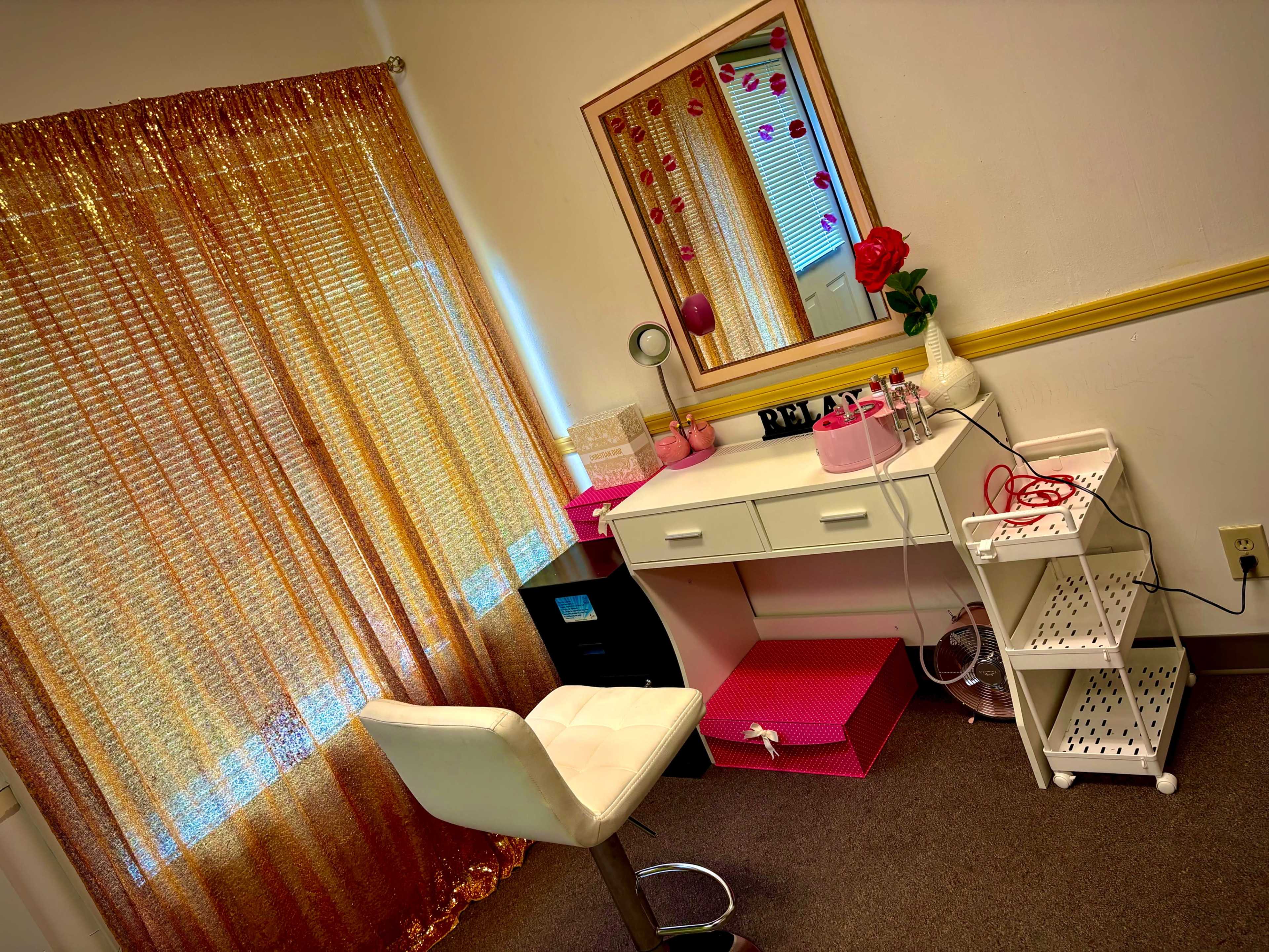 A vanity setup features a white desk with a mirror, a modern chair, and organized storage, accompanied by a decorative rose and curtains that filter light.