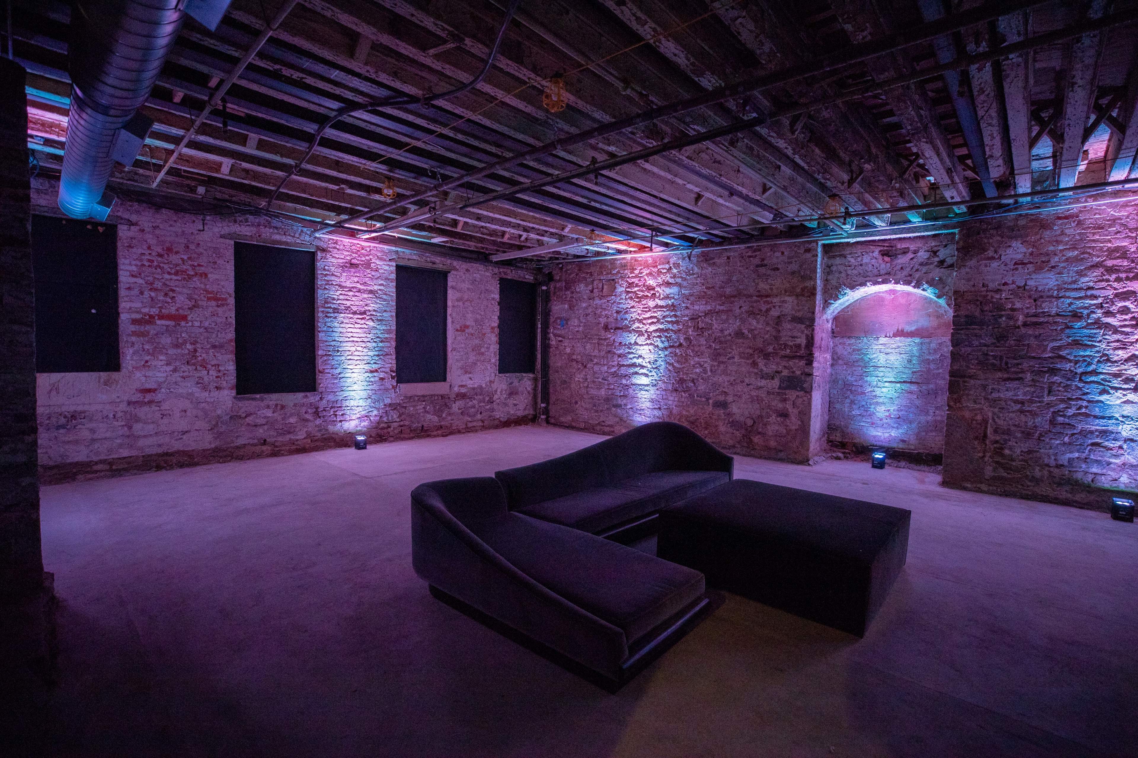 The image shows a dimly lit basement room with exposed brick walls and colorful lighting, featuring a modern black sofa set.